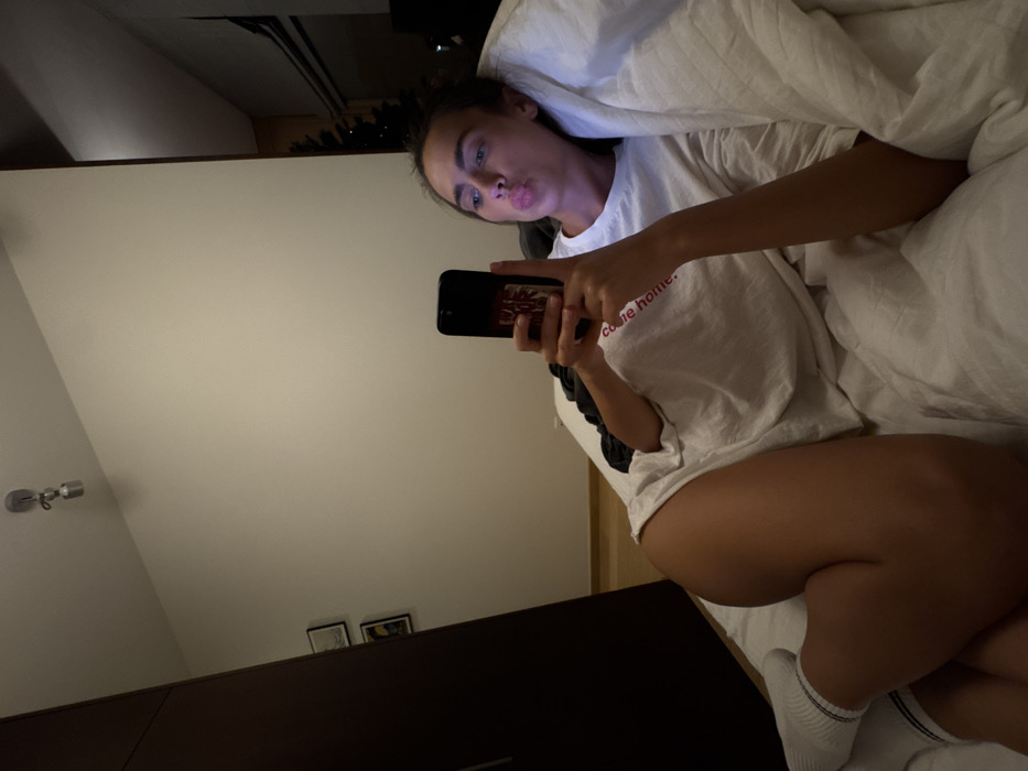 A woman in bed takes a selfie, her lips puckered. Partially covered by white bedding, she wears a white t-shirt with a short sleeve, dark colored detail, and red cursive text “come home.” Her phone case shows a red patterned design. One leg is visible, bent with a white sock on her foot. A wall with two small framed pictures and a light fixture can be seen. A dark colored cabinet or drawer unit is in the foreground.