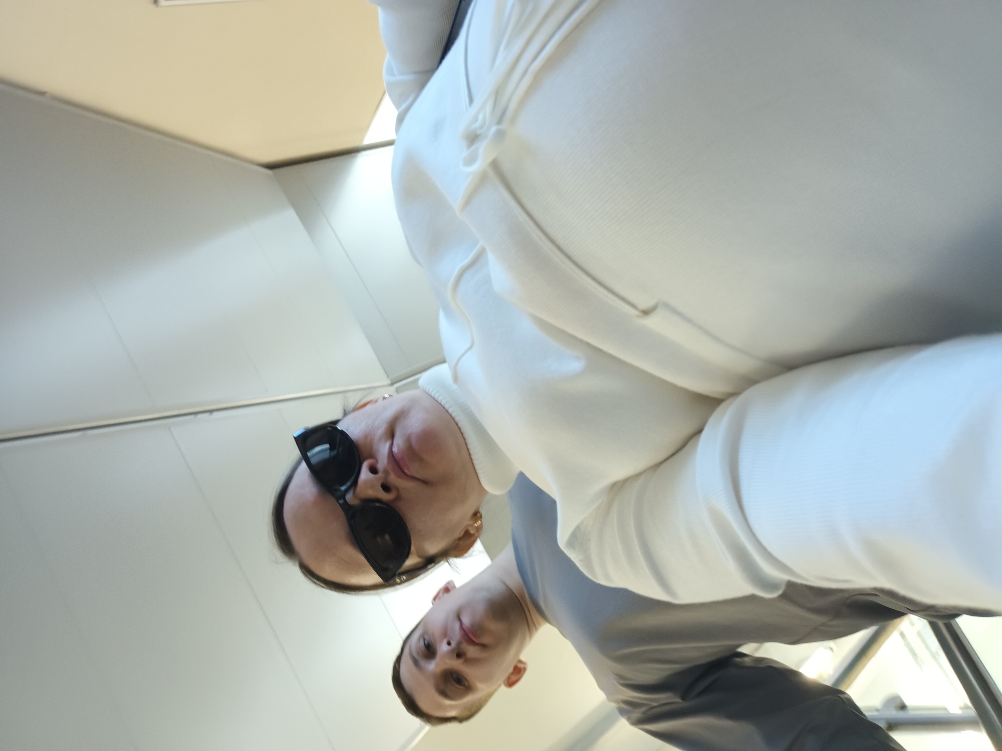 A photograph captures two people inside a structure featuring white walls and a cream-colored ceiling. The image is taken from a low angle, looking upwards at the subjects.
One individual, positioned closer to the camera, is wearing black sunglasses. They are dressed in a white sweater or sweatshirt. The other person, slightly behind and to the side, is wearing a gray shirt.
The location is Chaikovsky, Russia.