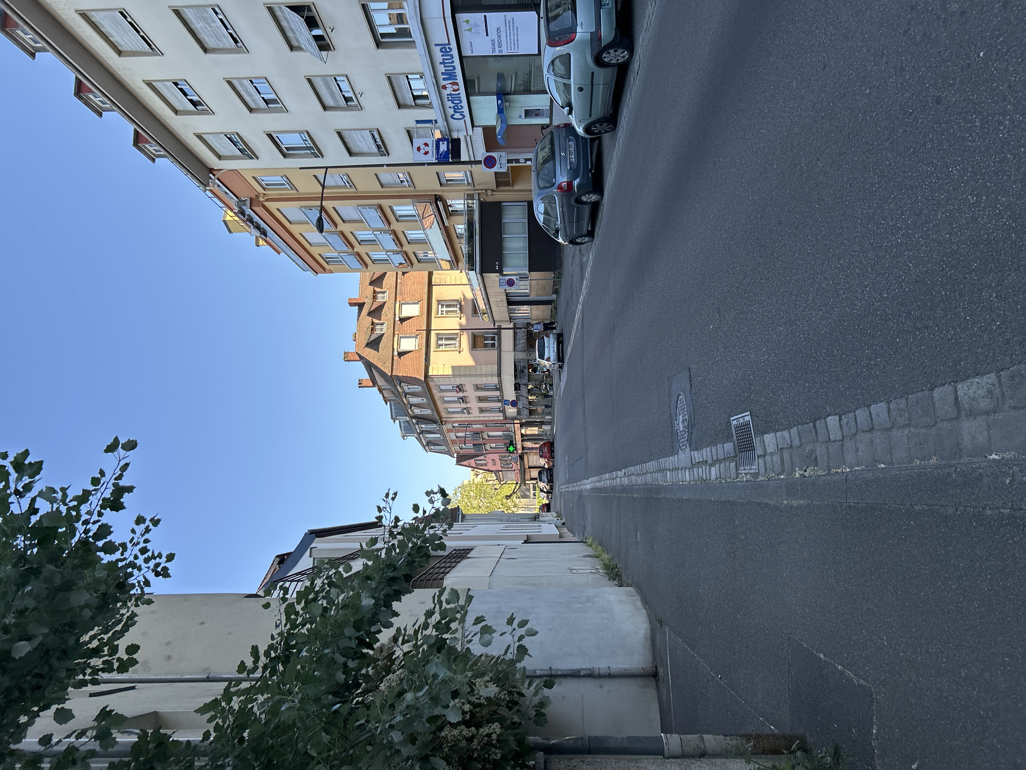 The image depicts a quiet urban street scene, likely in a European city given the architecture and visible text, confirmed to be Strasbourg, France. The perspective is from the side of the road, looking down the street. The sky is bright and clear blue, indicating sunny weather. The time of day appears to be either morning or late afternoon, as sunlight illuminates the building facades on the right side of the street and casts distinct shadows.
On the right side of the image, a row of multi-story buildings lines the street. The closest building has a light-colored facade with numerous windows, many of which have half-open or fully closed external blinds. Further down, buildings with varying architectural styles and colors, including warm orange and brick hues, are visible. Some buildings feature balconies.
The street itself is asphalt with a paved sidewalk bordering the buildings. Several cars are parked along the sidewalk on the right. A blue hatchback with a license plate "CM 880 AL" is prominent, next to a light blue or silver smaller car. A "Crédit Mutuel" bank branch is visible on the ground floor of one of the buildings, with its blue and red logo. A sign nearby states "TRAVAUX DE RÉNOVATION" (renovation work). A red circular sign with a blue border and a diagonal line, indicating "INTERDIT DE STATIONNER" (no parking), is also visible.
On the left side of the street, closer to the viewer, the foreground is dominated by green foliage from trees and bushes, obscuring parts of low-rise buildings or walls. Further down this side, more buildings are visible. The road surface includes manhole covers and grates. No people or overt activity are central to the frame; the scene is largely static, capturing the everyday street environment.