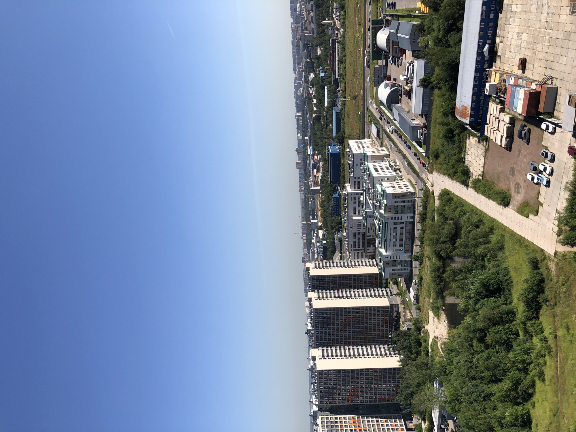 An aerial shot captures a bright, clear day with a vast expanse of blue sky dominating the upper portion of the image. Below, a cityscape unfolds with distinct zones. On the left, a cluster of tall, modern residential buildings with numerous windows are visible. These buildings are light-colored with dark facades and balconies. Further to the right, a more mixed urban landscape emerges, featuring lower-rise, white and green buildings that appear to be commercial or office spaces, surrounded by green areas and trees. A road with several cars parked and in motion winds through this section. In the foreground and mid-ground, industrial buildings are evident, including large, grey structures with curved roofs and a long, blue building with multiple windows. The ground in front of some of these buildings is a paved lot covered with stacks of concrete blocks, shipping containers, and several cars. A small body of water, possibly a pond or lake, is nestled amongst lush green trees on the left side of the image, with a dirt path leading towards it. The overall scene depicts a developing urban environment with a mix of residential, commercial, and industrial areas under a clear blue sky.