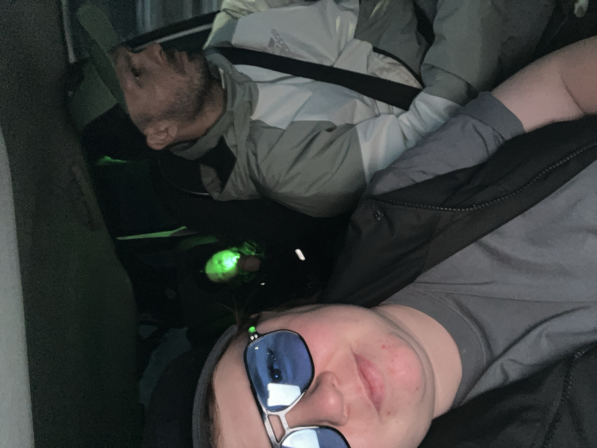 Two individuals are visible inside a vehicle. The person closest to the camera is wearing sunglasses and a grey top. The other person, further in the background, is wearing a jacket. A green light source is present in the middle of the scene. The location in Chaikovsky, Russia is not discernable.