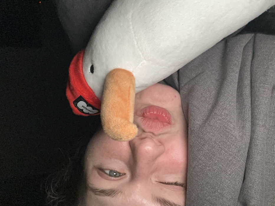 A close-up image depicts the left side of a person's face in direct proximity to a white plush goose toy. The person, with fair skin and a visible light-colored left eye (appearing blue or green), has their lips puckered forward, creating a "kissing" expression. Their right eye is mostly obscured or squinted shut. Dark, wavy hair is visible on the left side of the head. The plush toy, positioned to the right of the person's face, features an orange beak, a small black eye, and wears a red beanie with a black tag displaying Cyrillic text, "МИМИКЫШИ". The toy's beak is positioned near the person's puckered lips. Both the person and the toy are resting against a gray, textured fabric, likely a blanket or clothing. The background is dark, indicating a dimly lit indoor environment. The image itself does not provide visual cues to confirm a specific location within Kazan, Russia, or Russia generally.