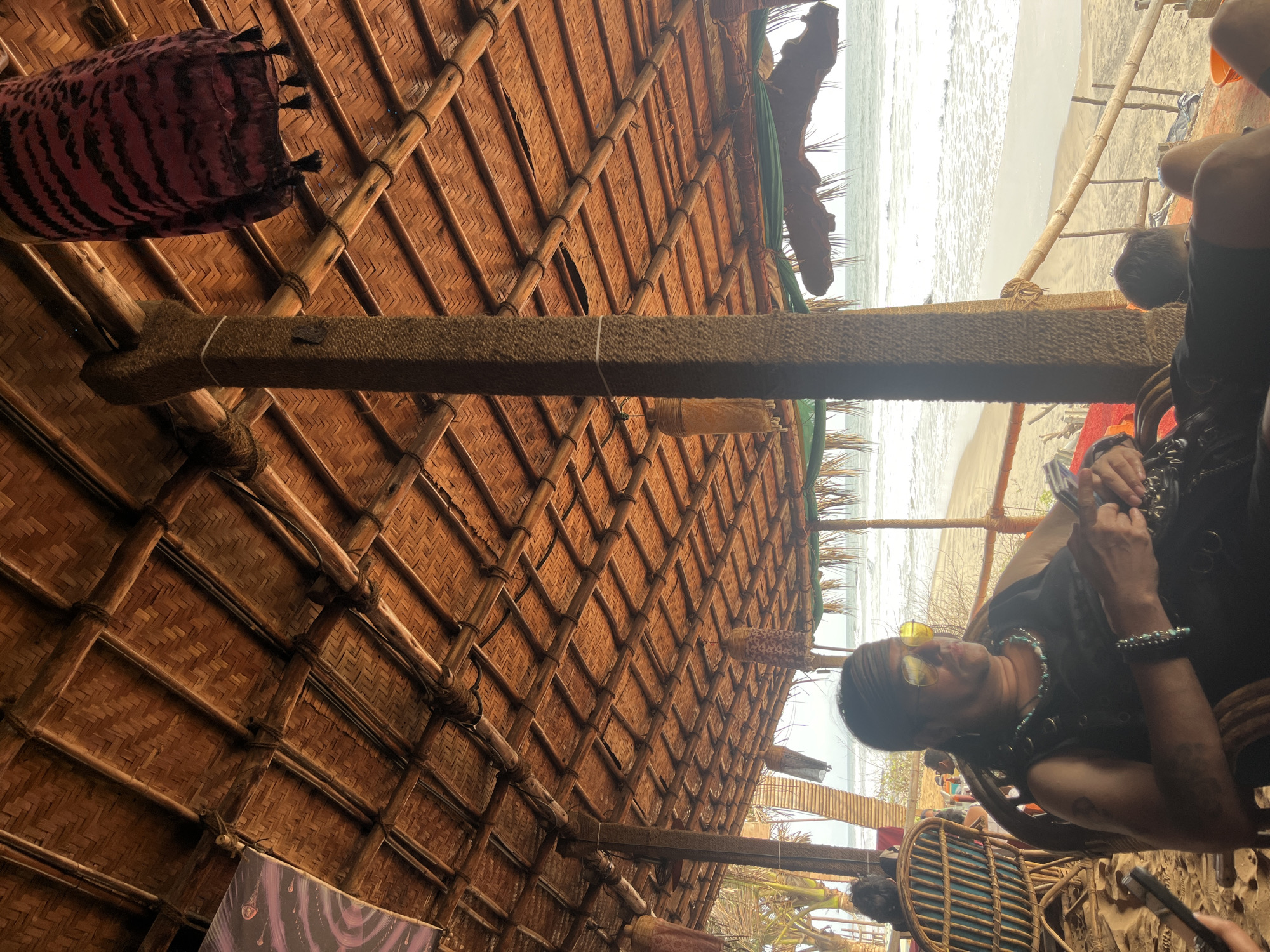 The image captures a scene at Ashvem Beach in India. The view is partially obstructed by the thatched roof of a beach hut, constructed with a bamboo frame. Various decorative fabrics hang from the roof's interior. Through the roof's opening, the sandy beach and ocean are visible. A person wearing sunglasses, a dark shirt, and jewelry sits in a wicker chair within the hut. They appear to be looking at or interacting with a mobile phone in their hands. Parts of other seated individuals are also visible in the background.