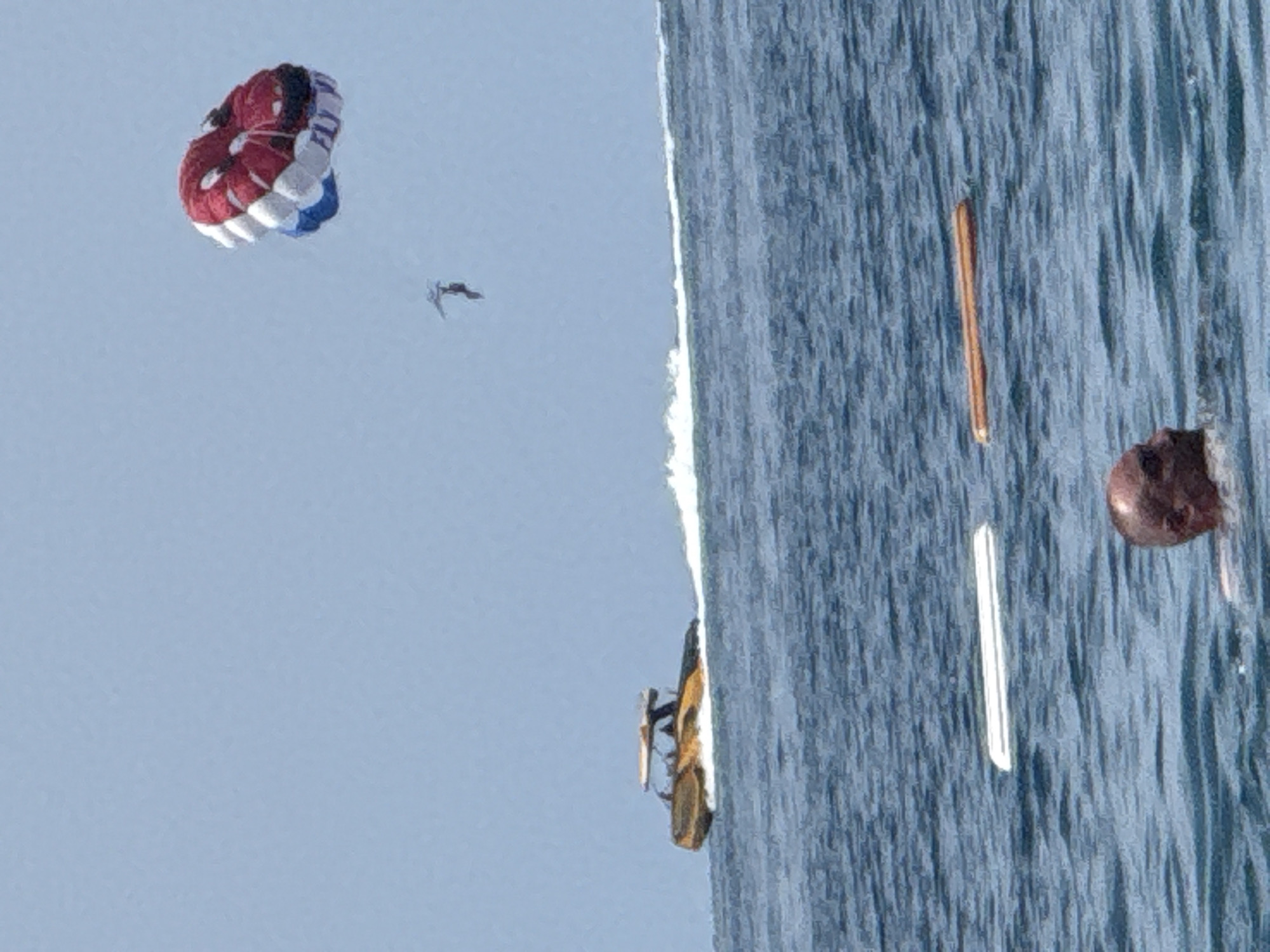 In the waters near Boreti, Montenegro, a parasailer is airborne, tethered to a speeding boat that is generating a white wake. A red, white, and blue parasail is visible in the sky above, with a person suspended beneath it. In the foreground, a person's head is visible above the water, and two floating objects, one white and one orange, are present in the sea. The sky is clear and blue.