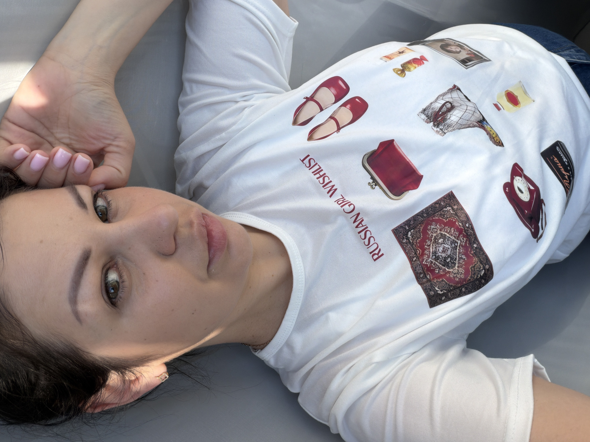 A woman is lying down, looking up at the camera. She has dark hair pulled back and is wearing a white t-shirt with a graphic print. The print features a collection of items, including a red purse, red shoes, a rug, a boot, a bottle of perfume, a framed picture of a woman, a rotary phone, a box, and a bottle. The woman is wearing a small gold earring. Her right hand is resting on her forehead, and her fingernails are painted pink. The background appears to be a flat, neutral surface. The lighting suggests the scene is outdoors, possibly during the day. There is no indication of the city of Sochi, Russia, in the image.