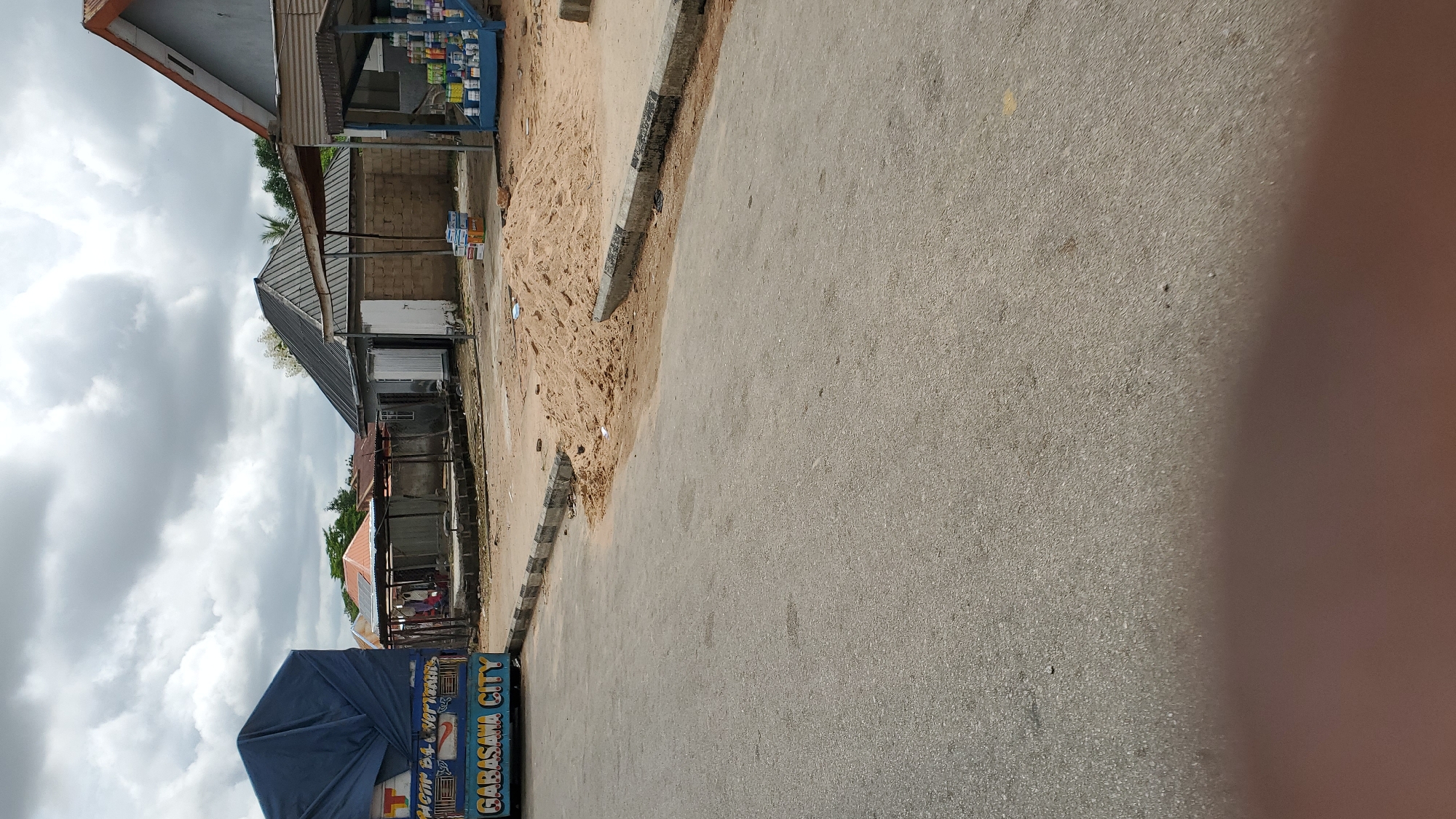 The image depicts a street scene in Jalingo, Nigeria, under a cloudy sky. In the foreground, a paved road occupies the majority of the frame. On the left, the rear portion of a blue tarp-covered truck is visible, with lettering on its side that reads "GABASANA CITY". Adjacent to the truck, there are some roadside stalls or shops with corrugated metal roofs. In the middle ground, a pile of sand is situated by the roadside, next to a concrete curb. Behind the sandpile and stalls, there are several buildings with metal roofs, some appearing to be residences or small businesses. The overall environment suggests a typical urban or semi-urban setting in Nigeria.