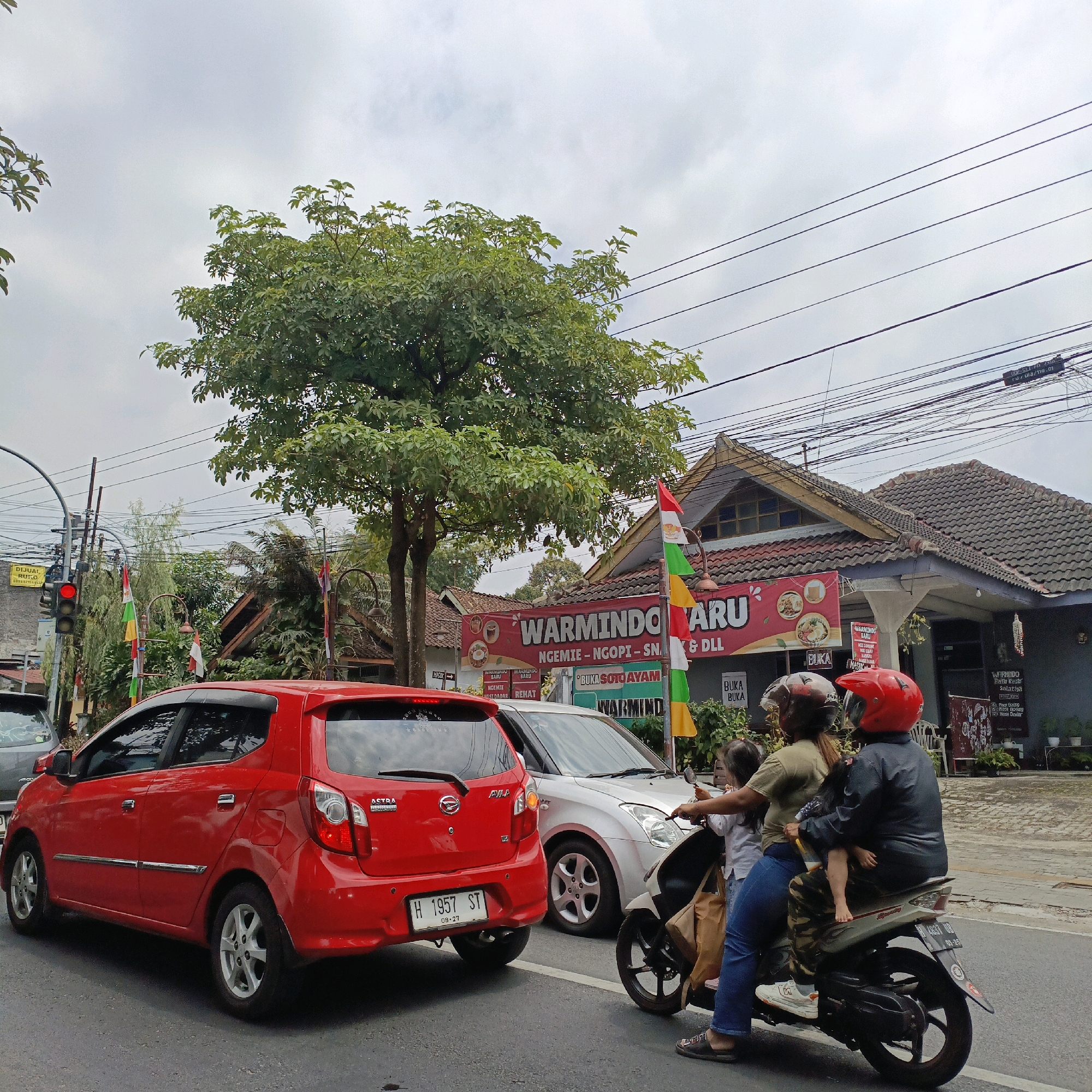 The image depicts a street scene in Salatiga, Indonesia, on an overcast day. In the foreground, a red car is stopped at what appears to be an intersection, indicated by a traffic light displaying red. A silver car is positioned to the right of the red car, and behind it, a scooter carries three people: an adult driver wearing a red helmet, an adult passenger in a dark jacket and helmet, and a child sitting in front of the passenger. The scooter is also stopped.
To the right of the vehicles, a building with a tiled roof and signs is visible. One prominent red banner reads "WARMINDORARU" with additional text below indicating food and drink offerings. Flags, including the Indonesian flag, are displayed on poles near the building. In the background, trees and power lines are present. The street surface is asphalt. The overall atmosphere suggests a pause in traffic flow, possibly due to the red light or congestion.
