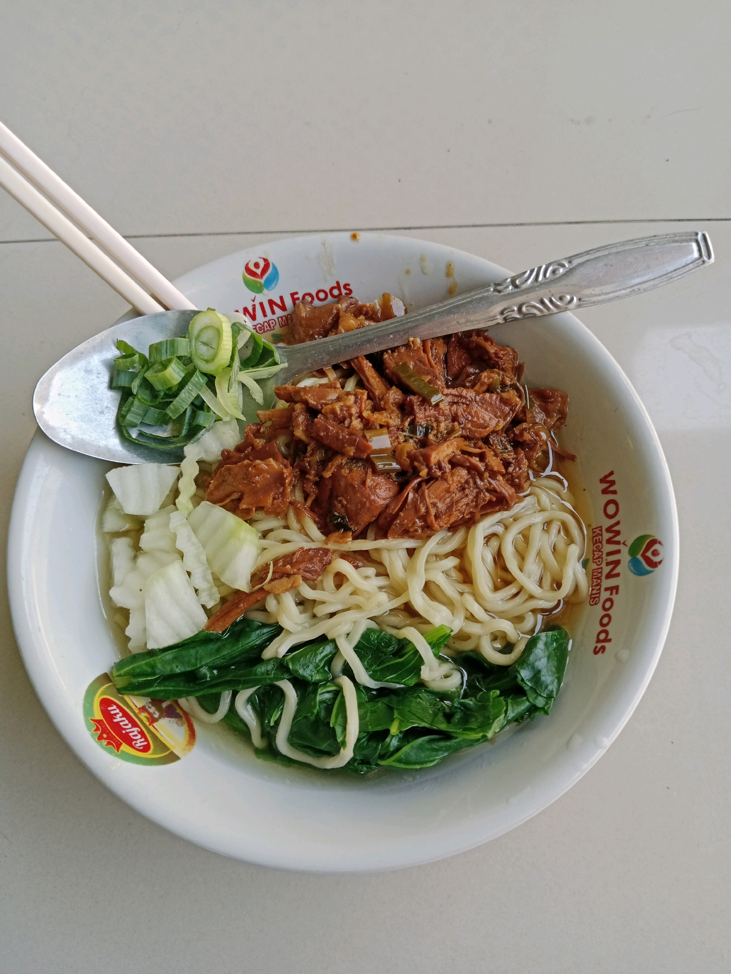 A close-up overhead shot shows a white bowl filled with chicken noodles and a side of vegetables. The bowl is white with red lettering that reads "WOWIN Foods KECAP MANIS". Inside the bowl, there are noodles, shredded chicken, chopped green onions, chopped white vegetables, and some leafy green vegetables. A spoon and a pair of chopsticks are placed on top of the bowl. The setting appears to be a table in Karanganyar, Indonesia, with a light-colored tiled surface visible in the background.