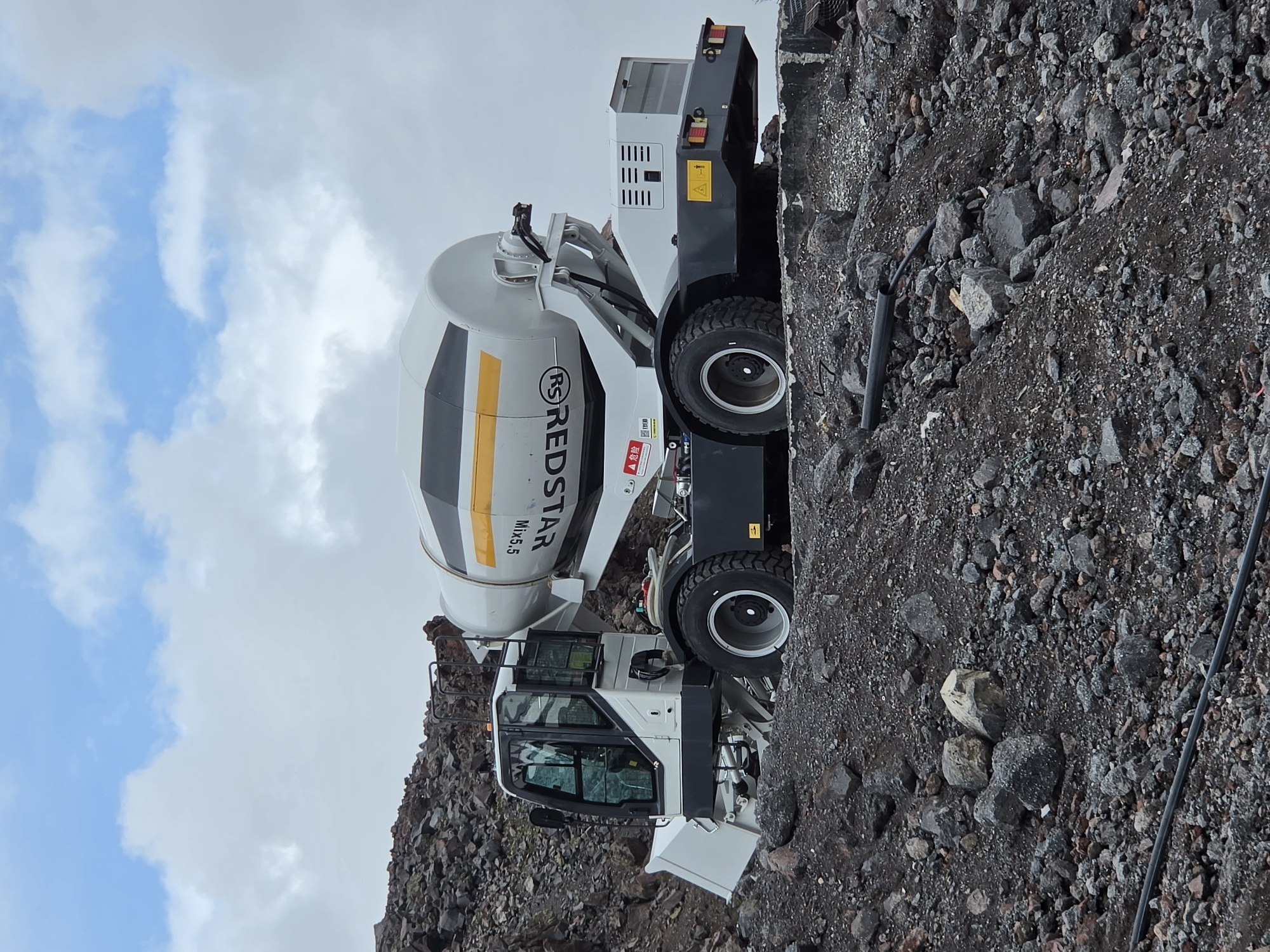 A white and grey concrete mixer truck is parked on a pile of dark, rocky debris. The truck has a large cylindrical drum on the back, painted with yellow and black stripes and the brand name "REDSTAR" in yellow letters. The truck's cabin is white with large glass windows. The scene appears to be an outdoor construction or demolition site, characterized by the rough terrain and scattered rocks. The sky above is cloudy with patches of blue. The location of Terskol, Russia, is indicated as the context for this image. There are no people visible in the image.