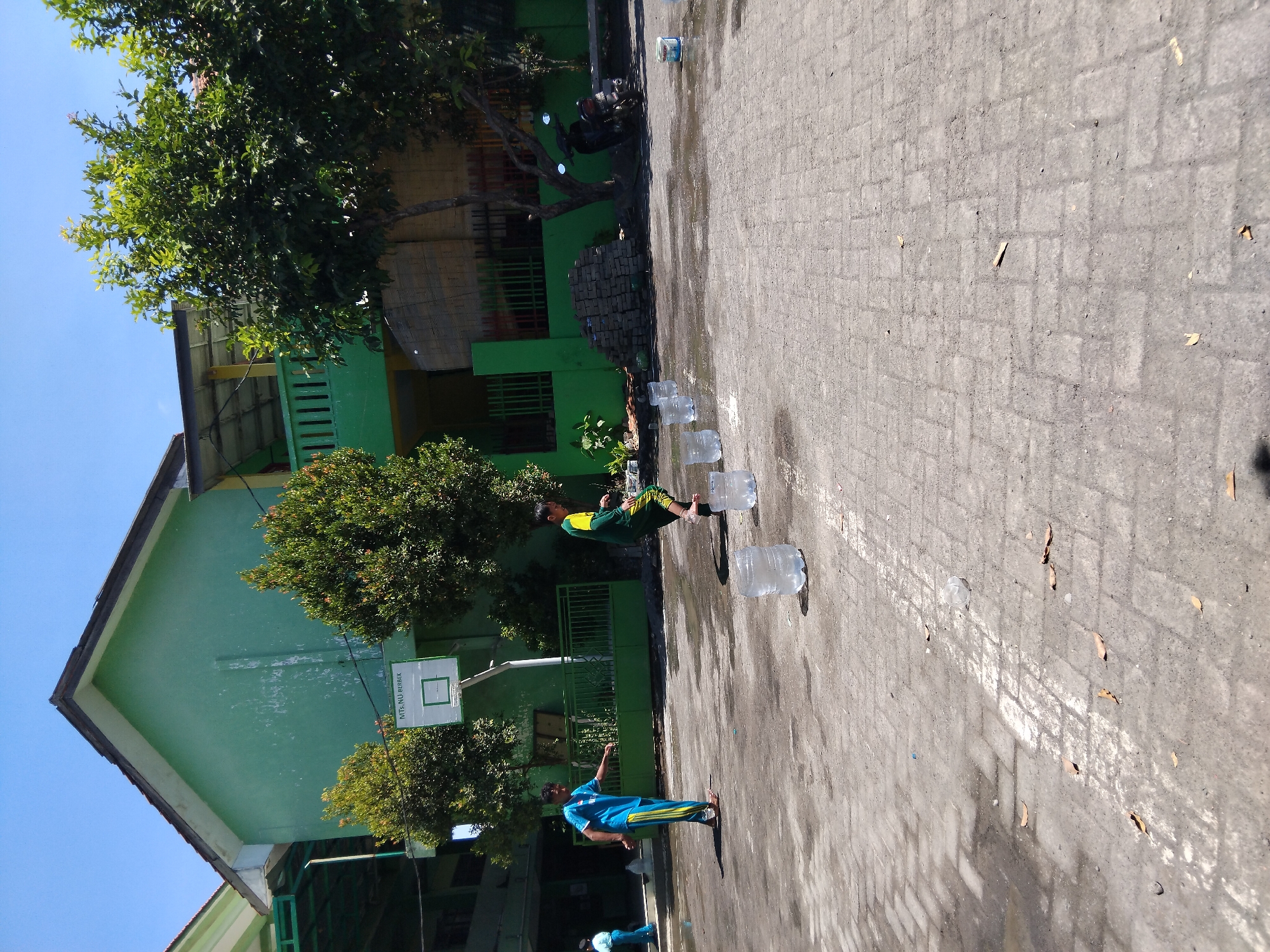 A bright, sunny day with a clear blue sky over an outdoor concrete courtyard in Kepuh Kiriman, Indonesia. The courtyard is paved with interlocking gray bricks, and there are scattered puddles of water reflecting the sunlight. On the left side of the frame, a two-story building with a green facade and a brown roof is visible. A basketball hoop is mounted on the green wall. Trees with green foliage grow in front of the building. Two boys are playing in the courtyard. One boy, wearing a blue shirt and yellow and blue striped pants, stands with his left leg forward, his arms extended, and his head turned to his right. The second boy, wearing a green shirt with yellow sleeves and green pants, is in motion, stepping with his right foot onto a clear plastic water bottle. There are several more clear plastic water bottles placed in a line across the courtyard, appearing to serve as stepping stones. A pile of gray bricks and a dark-colored motorcycle are visible near the building on the right.