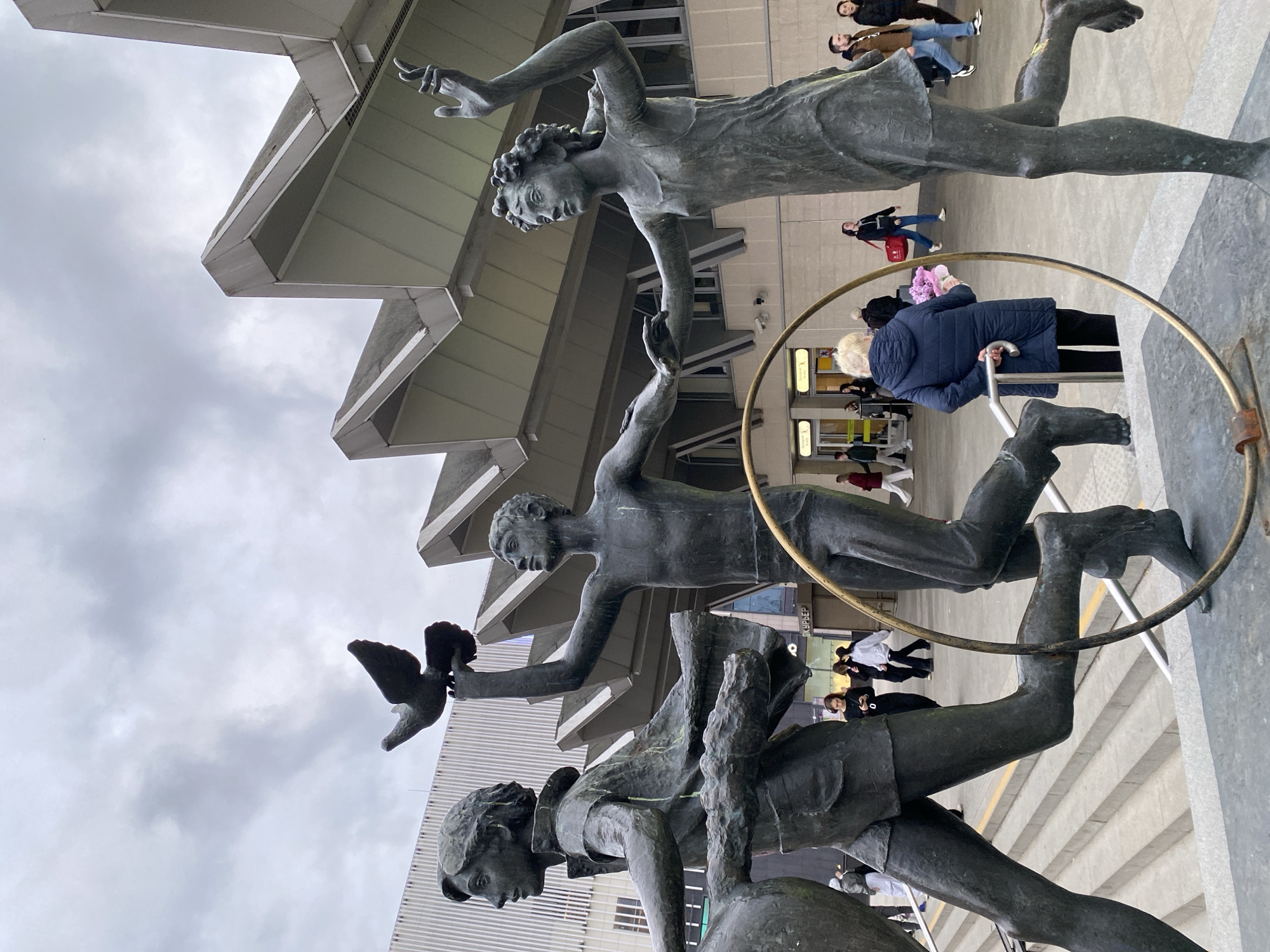 The image captures a bronze statue of three figures in motion outside the Moskovskaya Metro Station in Saint Petersburg, Russia. The figures appear to be soaring or running, with one holding a bird. A large bronze hoop encircles them. People are seen walking by in the background, some entering or exiting the station, and a modern building with a stepped facade stands behind the statue. The weather appears overcast with a cloudy sky.