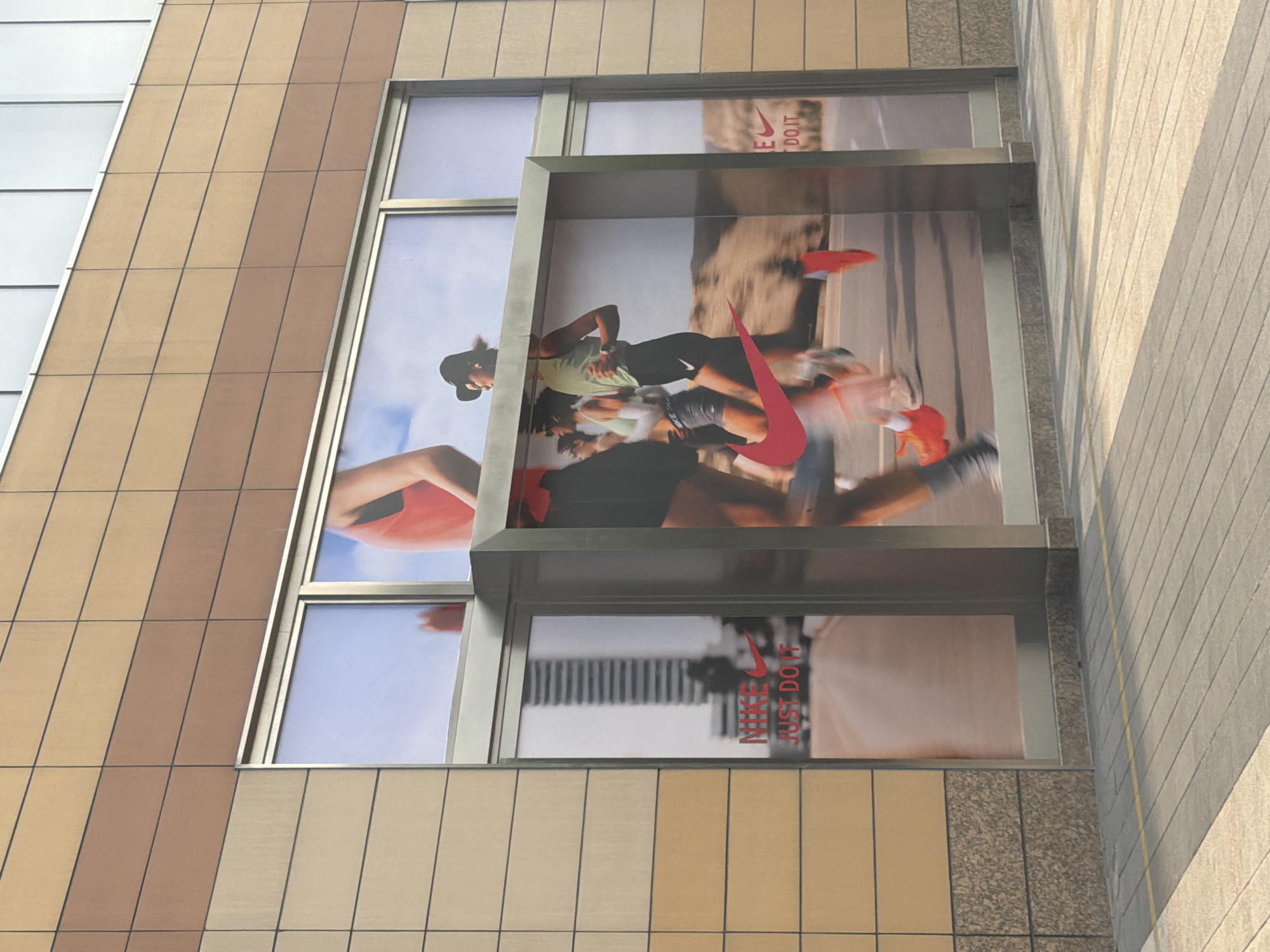 This image captures a section of a modern building facade in Yuhuatai District, China, featuring large advertising displays. The building's exterior is constructed with alternating bands of light beige and brown rectangular panels, arranged in a grid pattern.
Prominently featured are multiple window displays showcasing a Nike advertisement. The main display depicts several people in athletic wear engaged in running. A central figure, a woman wearing a light-colored top, dark shorts, and a cap, is shown mid-stride with other blurred figures around her, conveying a sense of dynamic motion. A large, bold red Nike swoosh logo is superimposed over the runners.
Visible text within the advertisements includes "NIKE" and its iconic swoosh, as well as the slogan "JUST DO IT," both rendered in red against the background imagery of the runners and a bright sky. These elements appear in both the larger central display and smaller adjacent window sections.
The scene is brightly lit, suggesting it was captured during daylight hours. The architectural style is contemporary, characterized by clean lines and mixed materials like glass, metal, and composite panels. The overall impression is that of an urban commercial environment.