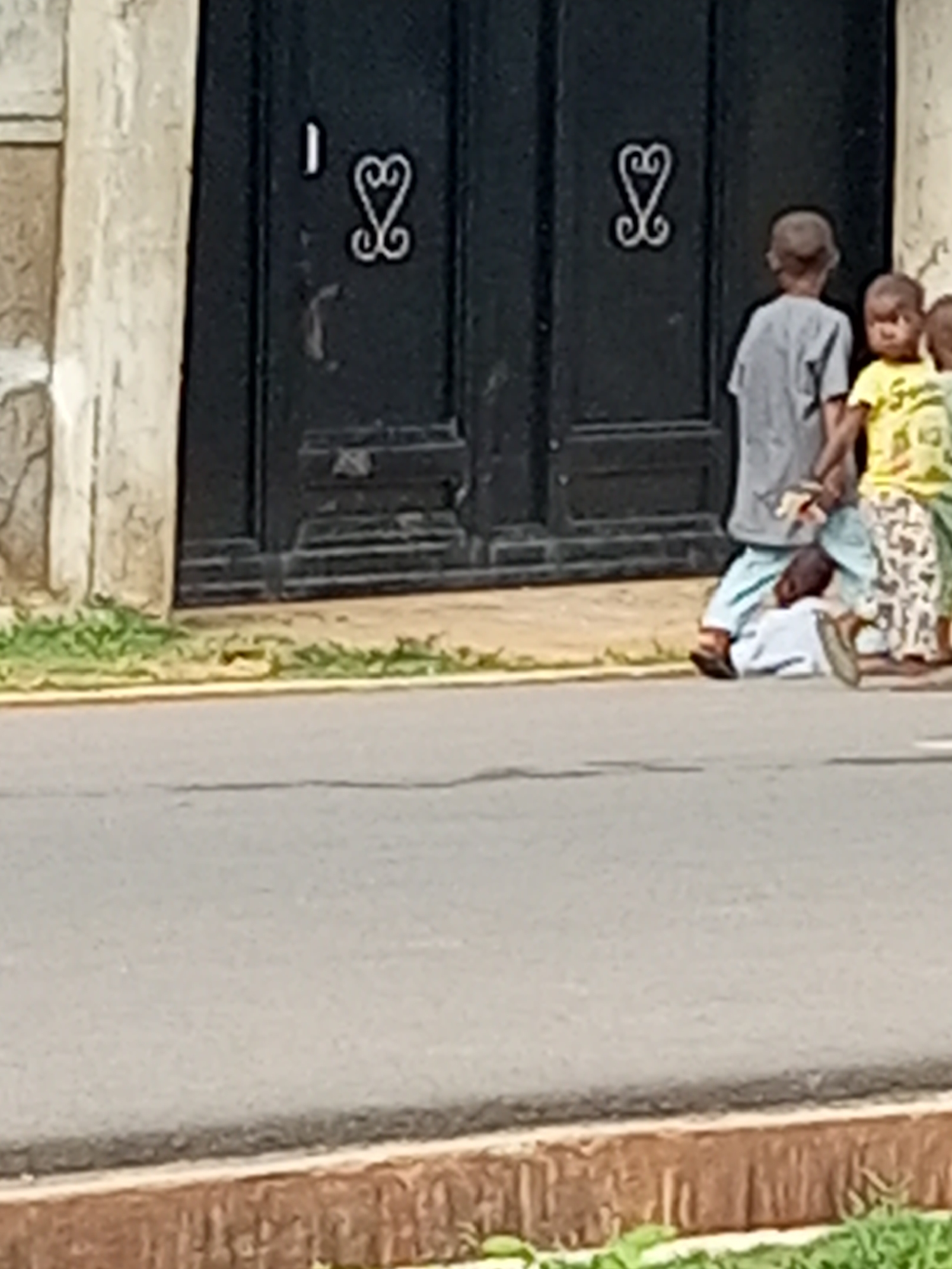 In front of a dark double door featuring decorative metal heart shapes, two young children stand near a third child lying on the ground. The child on the ground is wearing a light blue garment. One standing child, wearing a grey t-shirt, is turned away from the camera. The other standing child wears a yellow t-shirt and is facing the camera. The scene is on a paved surface, suggesting a street or pathway, with a strip of green grass and a yellow border separating it from the adjacent area. The buildings in the background appear to be made of concrete.