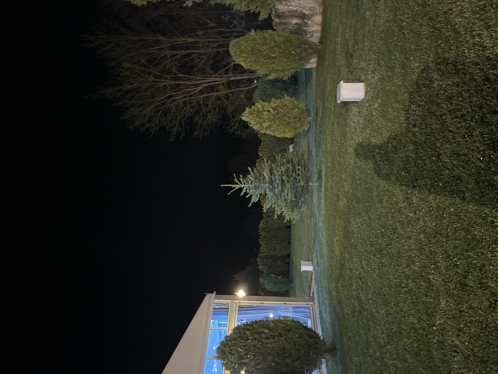 A nighttime view shows a lawn in Koprivlen, Bulgaria. Trees, including bare branches and conifers, border the lawn. A building with illuminated windows stands at the edge of the frame. White rectangular objects are placed on the lawn. The scene is dark with artificial lighting illuminating some parts.