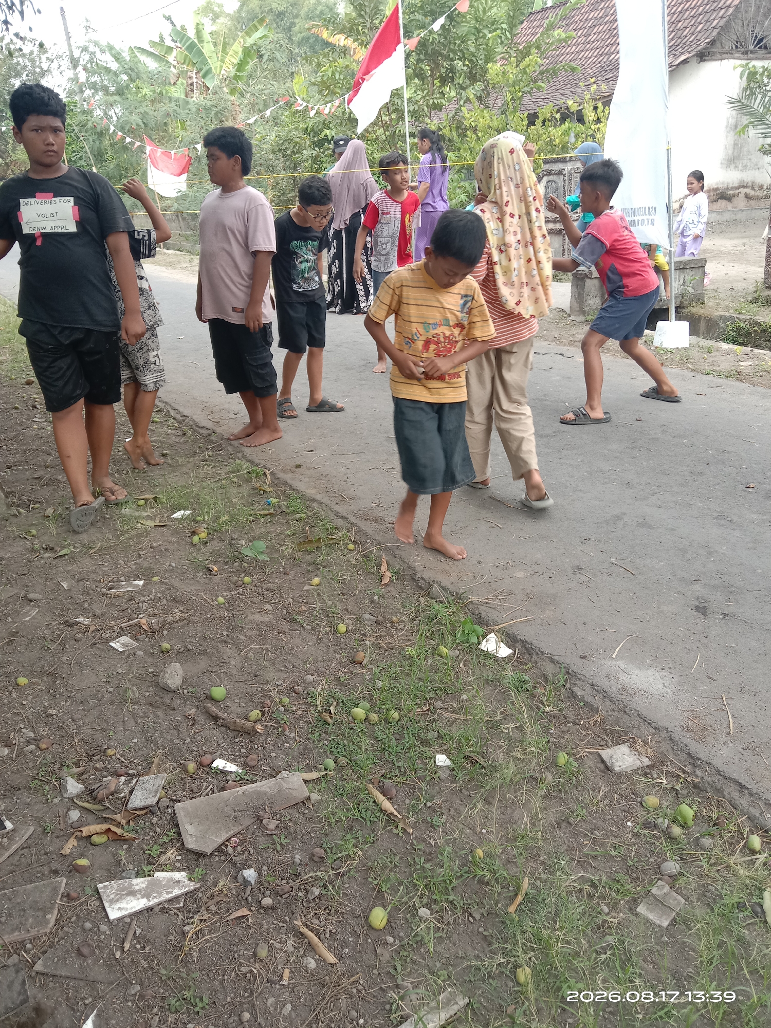 In Banyuarang, Indonesia, several children are gathered on a paved road, with a dirt and grassy area to the left. The children appear to be engaged in a game or activity. One boy in a black t-shirt with white lettering is in the foreground. To his right, another boy in a pink t-shirt stands. Further down the road, a boy in a black t-shirt with a graphic design and glasses is visible. A child in a red and blue t-shirt and blue shorts is playing. Several other children are present in the background, some with adult supervision, and flags and decorations suggest a local celebration. The environment is rural, with trees and vegetation bordering the road, and some debris scattered on the ground. The date stamp on the image indicates August 17, 2026, at 1:39 PM.
