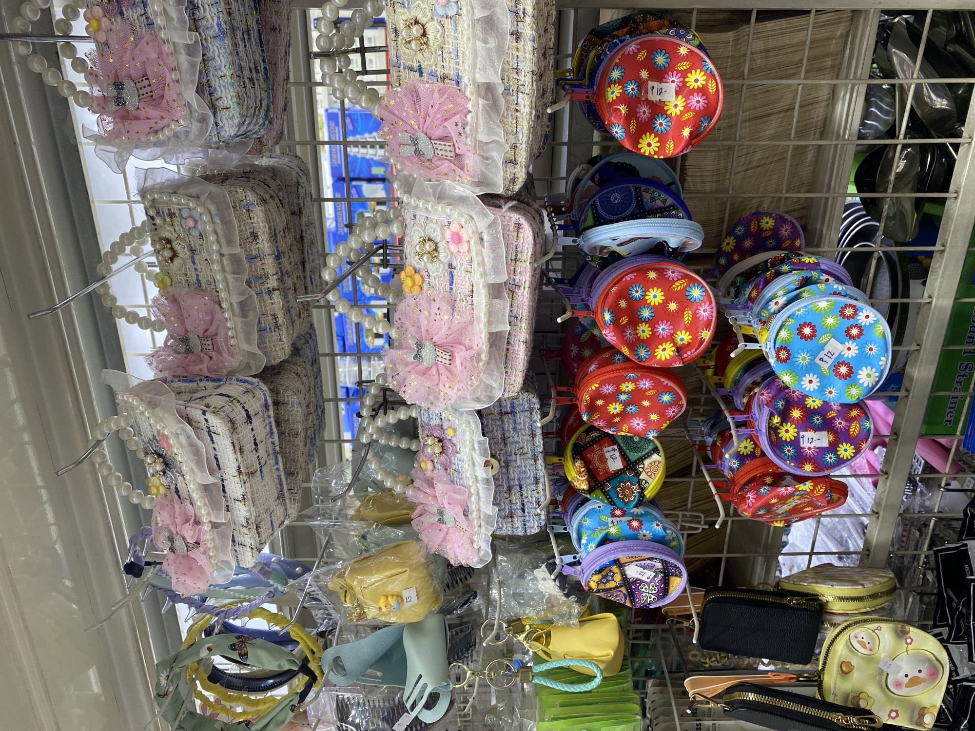 The image shows a retail display of various small bags and accessories, likely in a store. In the foreground, rows of colorful, round coin purses with floral and patterned designs are hung on a metal rack. Some of these purses have price tags attached. Below these, a yellow coin purse with a snowman design is visible. To the left, several black and brown zippered pouches are also displayed. In the midground, a collection of small, decorative handbags with pearl handles, lace trim, and pink floral embellishments are hung on a higher rack. These bags appear to be made of a textured, woven fabric. Further up and to the left, a selection of headbands with various colors and designs, including some with butterflies and bows, are displayed. Yellow and teal small pouch-like items are also visible on hooks. The background shows a blurry shelving unit with what appear to be packaged goods. The overall impression is a brightly lit retail environment with a focus on small, decorative items. The specific location is a store in Davao City, Philippines, possibly a department store or a specialty shop selling accessories.