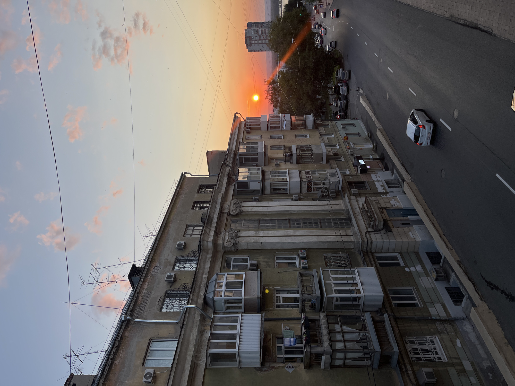 The photograph depicts a street scene in Samara, Russia, during sunset. The view is from an elevated position, looking down onto a multi-lane road lined with parked cars and moving vehicles. On the left, a weathered, multi-story apartment building dominates the foreground, its facade featuring balconies and numerous windows. The building's architecture suggests it is an older Soviet-era residential block.
The sky is a gradient of orange and pink, with scattered clouds, indicating the setting sun. The sun itself is a bright, small orb low on the horizon, casting a warm glow over the scene. In the distance, another tall apartment building is visible, and beyond that, a body of water, possibly the Volga River, can be seen.
The street appears to be a main thoroughfare, with white dashed lines marking the lanes. A white car is in motion, heading away from the viewer. The sidewalks are paved with bricks. The overall atmosphere is one of urban twilight, with the warm light of the setting sun contrasting with the aging concrete structures. There are no visible people in the immediate foreground.