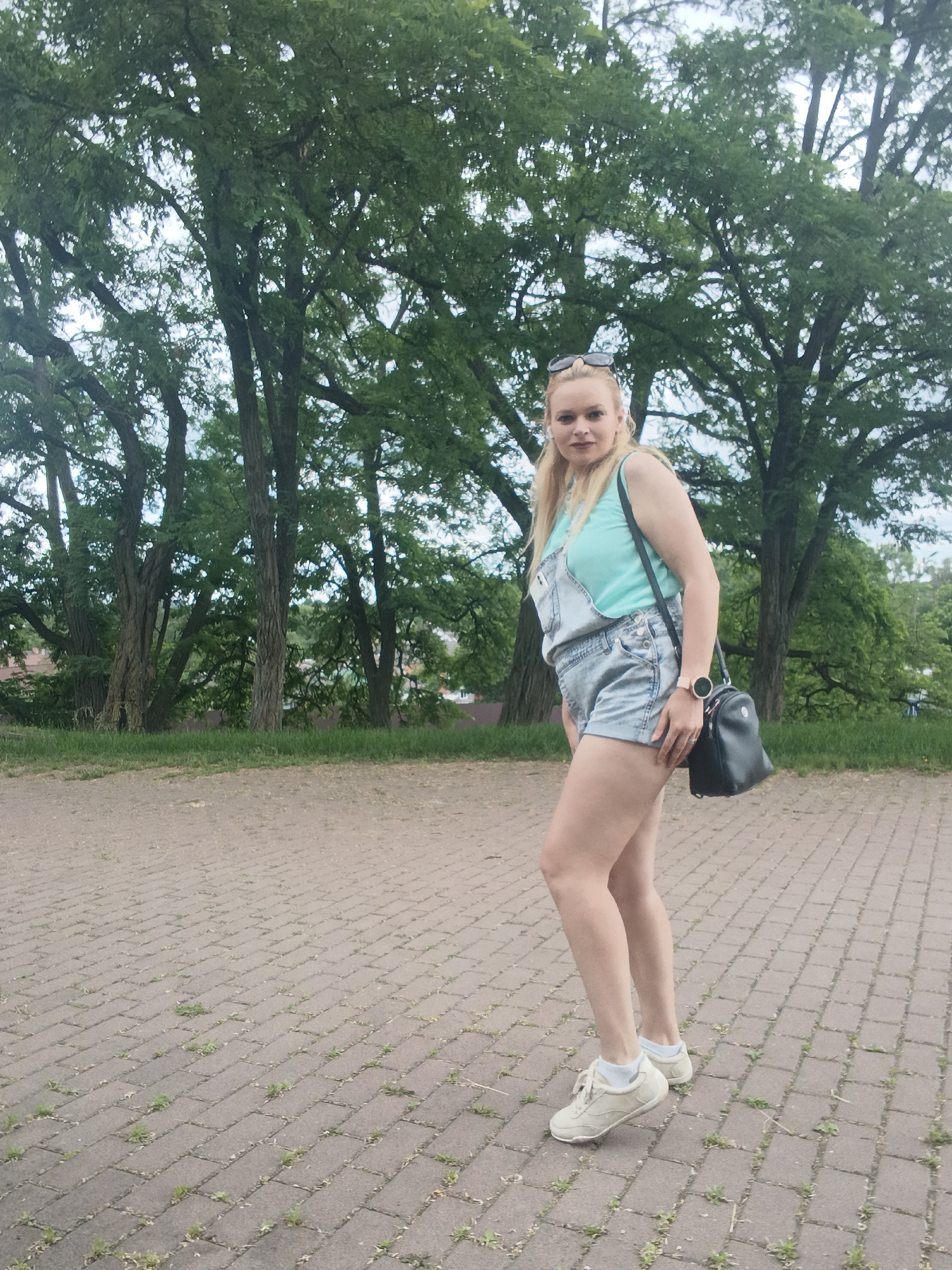 A blonde woman stands on a paved walkway in a park setting with trees and grass. She is wearing denim overalls, a teal tank top, and white sneakers. A black crossbody bag is slung over her shoulder, and sunglasses are pushed up on her head. The paved walkway is made of interlocking brickwork with sparse green weeds growing in the cracks. The background consists of large trees with dense green foliage, and a cloudy sky. The location is identified as Chernihiv, Ukraine.