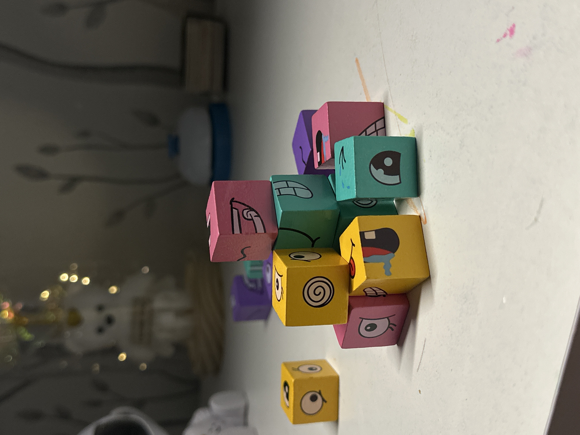 The image shows a collection of colorful wooden blocks with cartoon-like faces stacked on a white surface. The blocks are various colors including pink, purple, teal, and yellow. Each face is different, depicting various expressions like sadness, confusion, or happiness. The background is blurry, showing a dimly lit interior with other objects. Location information is not present.