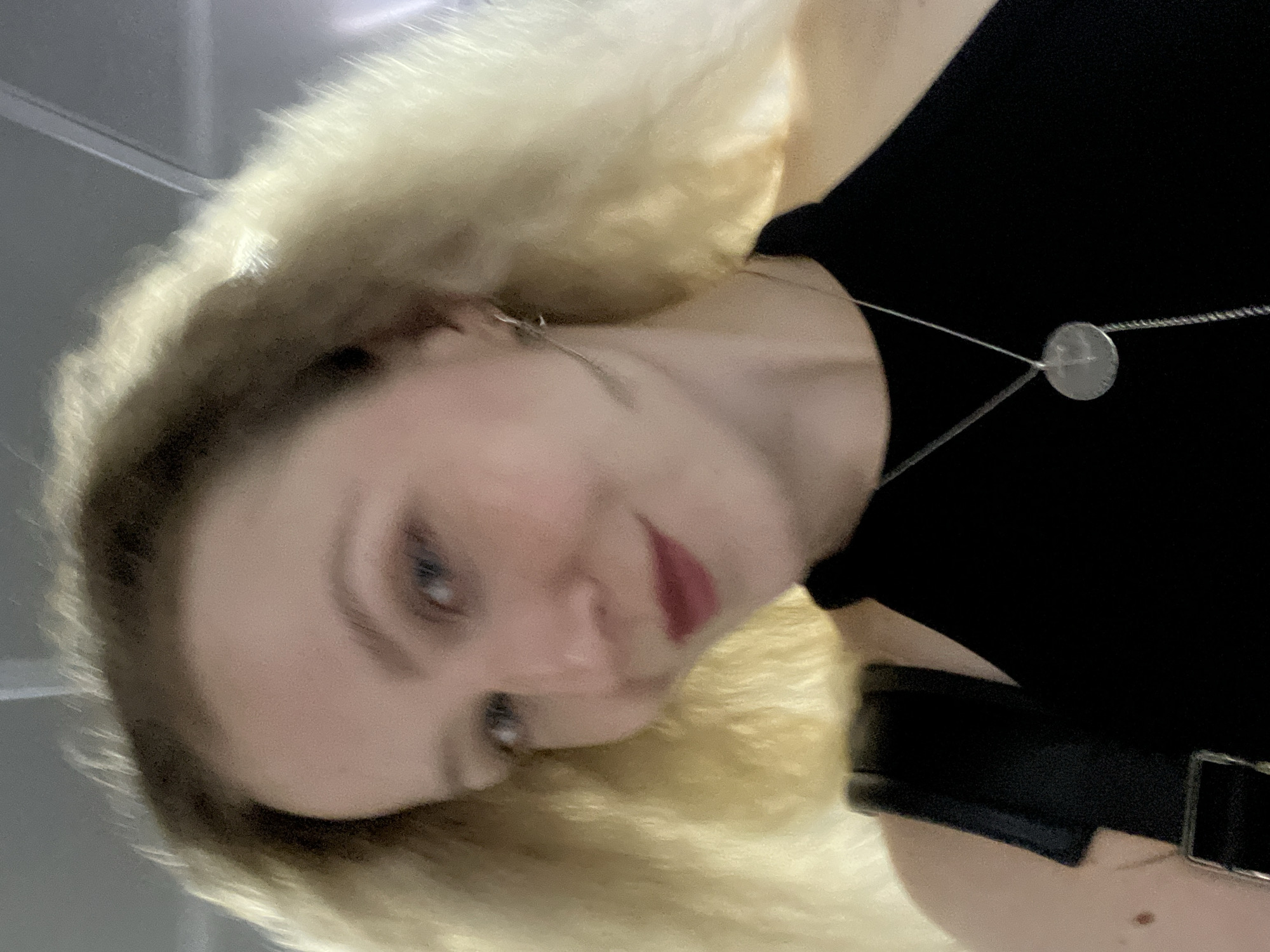 A woman with blonde hair is taking a selfie. She is wearing a black top and a silver necklace. The background appears to be indoors, possibly a public transit setting, given the grey ceiling and bright lighting which suggests a station or terminal. The image is taken from a low angle, looking up at the woman.