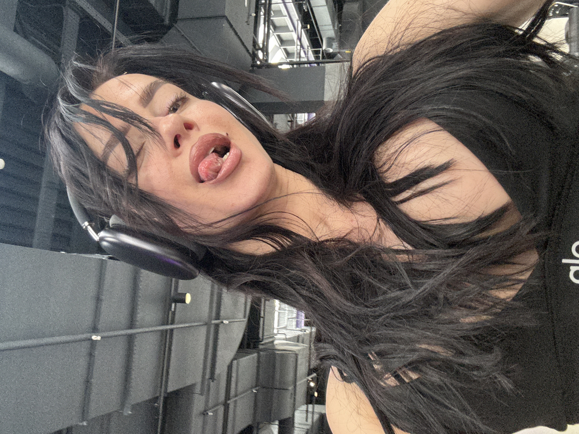 A woman with dark hair and headphones is posing with her tongue sticking out. She is wearing a black top with white lettering. The background appears to be an interior industrial setting with exposed pipes and ventilation systems. The exact location within the city of Krasnoyarsk, Russia is not identifiable from the image.