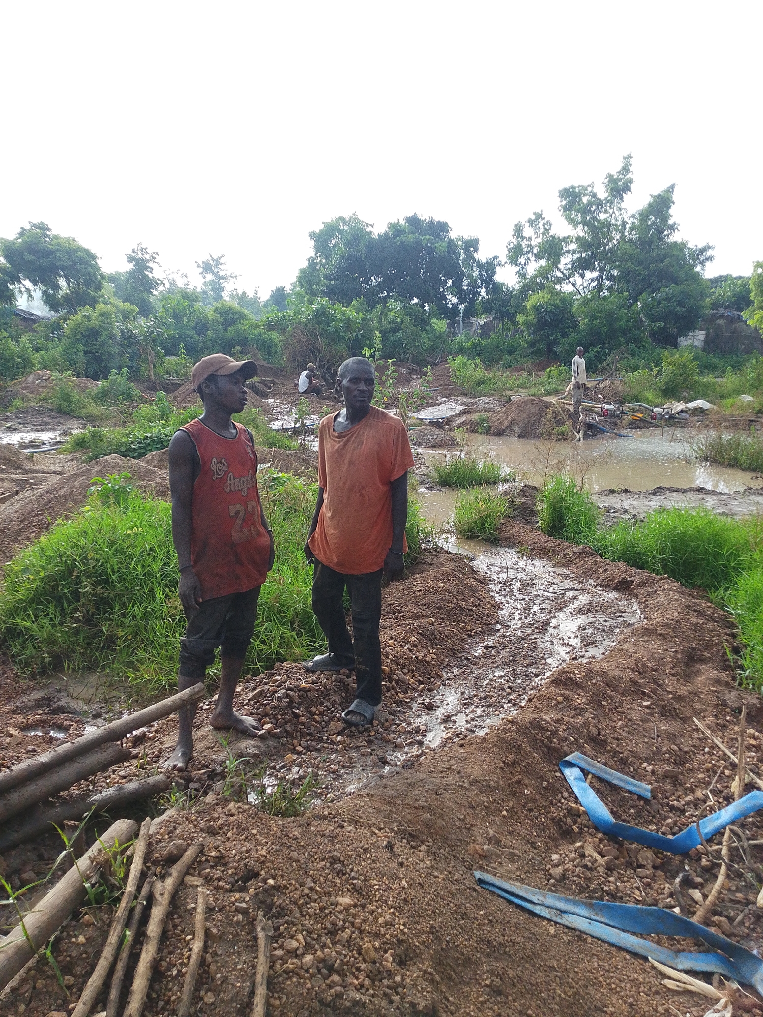 In Otulu, Nigeria, a scene unfolds at a waterlogged excavation site. Two men, their faces turned away from the camera, stand amidst piles of muddy earth and debris. The man on the left wears a red tank top with "Los Angeles 27" printed on it and a brown baseball cap. He has dark skin and appears to be in his late teens or early twenties. The man to his right, older and with darker skin, is dressed in an orange t-shirt and dark pants. His gaze is directed towards the muddy water and the equipment in the distance.
The environment is a chaotic landscape of human activity. The ground is a mix of dark, wet soil and scattered stones, with evidence of recent digging and earth movement. Patches of vibrant green grass struggle to grow through the disturbed earth. In the background, a murky body of water reflects the overcast sky. Several water pumps, likely for drainage or extraction, are visible near the water's edge, along with a large white bag, possibly containing materials. The overall impression is one of manual labor in a challenging, wet environment, with the lush vegetation providing a stark contrast to the disturbed earth.