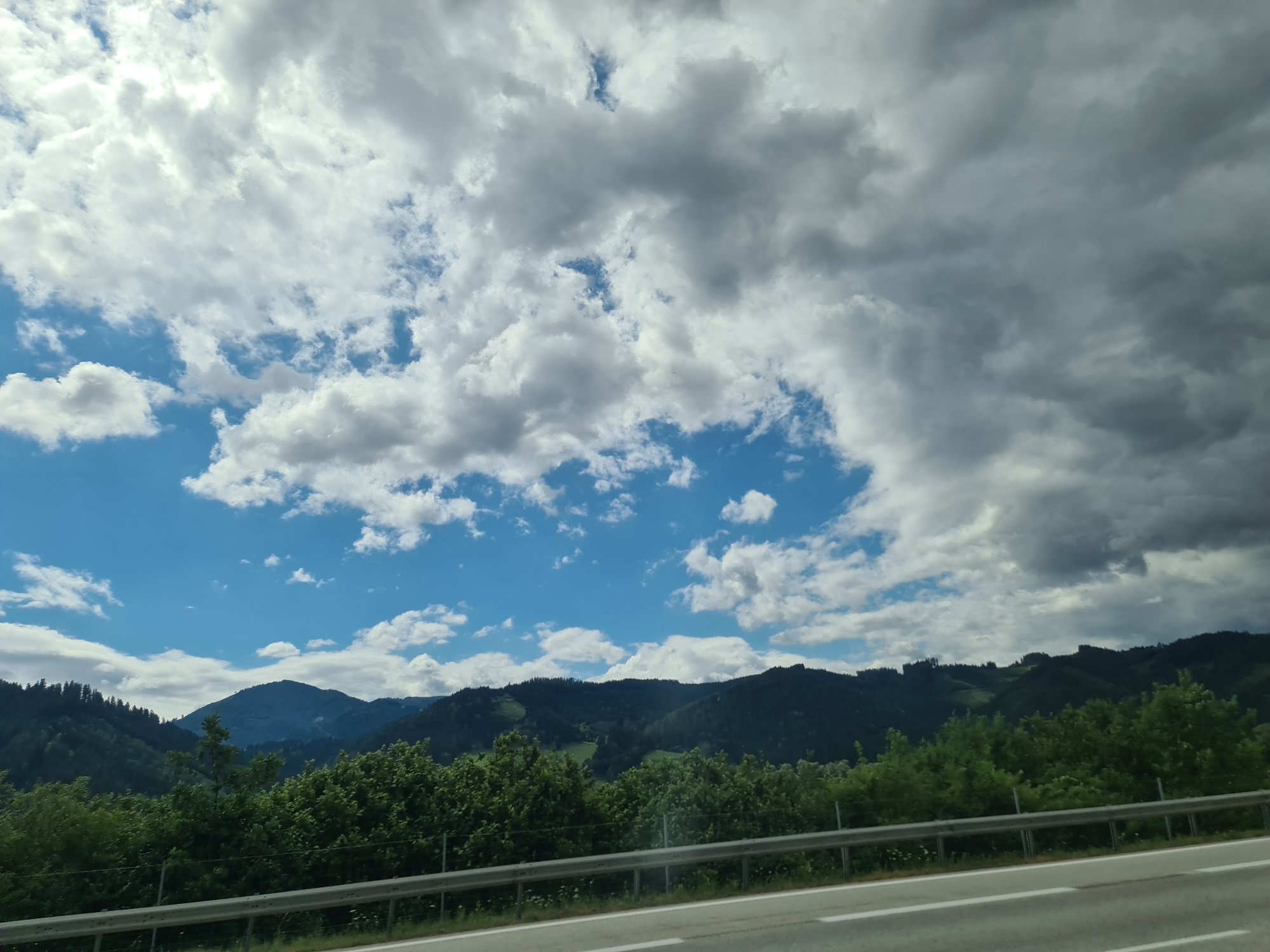The image captures a scenic view of a mountainous landscape under a cloudy sky. In the foreground, a road with white markings is visible, separated from the scenery by a silver guardrail. The middle ground is dominated by rolling green hills covered in dense trees, with some areas appearing as lush meadows or clearings. The background features several layers of dark, tree-covered mountains against a bright blue sky partially obscured by dramatic, varied cloud formations, ranging from bright white to dark gray. The lighting suggests it is daytime, with sunlight filtering through the clouds in some areas. The scene appears to be located in a rural or mountainous region, likely in Sankt Marein-Feistritz, Austria, given the characteristic Alpine scenery. There are no people or animals visible in the image.