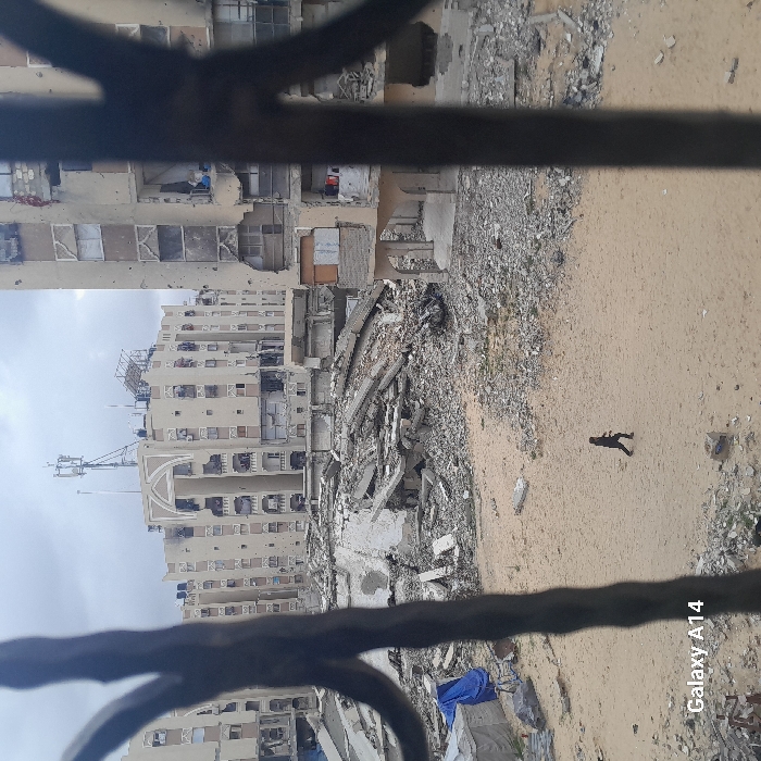 This image captures a scene of extensive urban destruction in Khan Yunis, Palestinian Territory, viewed from an elevated vantage point, partially framed by dark, vertical ironwork. The foreground and midground feature a desolate expanse of light-colored sand and widespread concrete rubble, rebar, and fragmented building materials.
In the lower midground, a lone individual, dressed in dark clothing, walks from left to right across the sandy terrain. Behind this figure, a massive, jagged pile of collapsed concrete slabs, twisted rebar, and debris dominates the center, indicative of a demolished structure. To the left of this rubble, a small, makeshift shelter covered with blue tarps is partially visible.
The background is filled with multi-story residential buildings, all exhibiting varying degrees of severe damage. The building directly behind the central rubble pile displays significant structural compromise, with large sections of its facade missing, exposing multiple interior rooms and showing areas of black scorching. To the right, another multi-story building features numerous impact holes across its exterior, fragmented walls, and shattered windows. Further back and slightly to the left, a larger, multi-story building with distinct arched architectural elements and multiple balconies also shows damage to its facade and windows, though it appears more structurally coherent than the immediate foreground buildings. The rooftops of these background structures are equipped with multiple black water tanks and communication towers/antennae. The sky above is overcast with a uniform grey cloud cover.