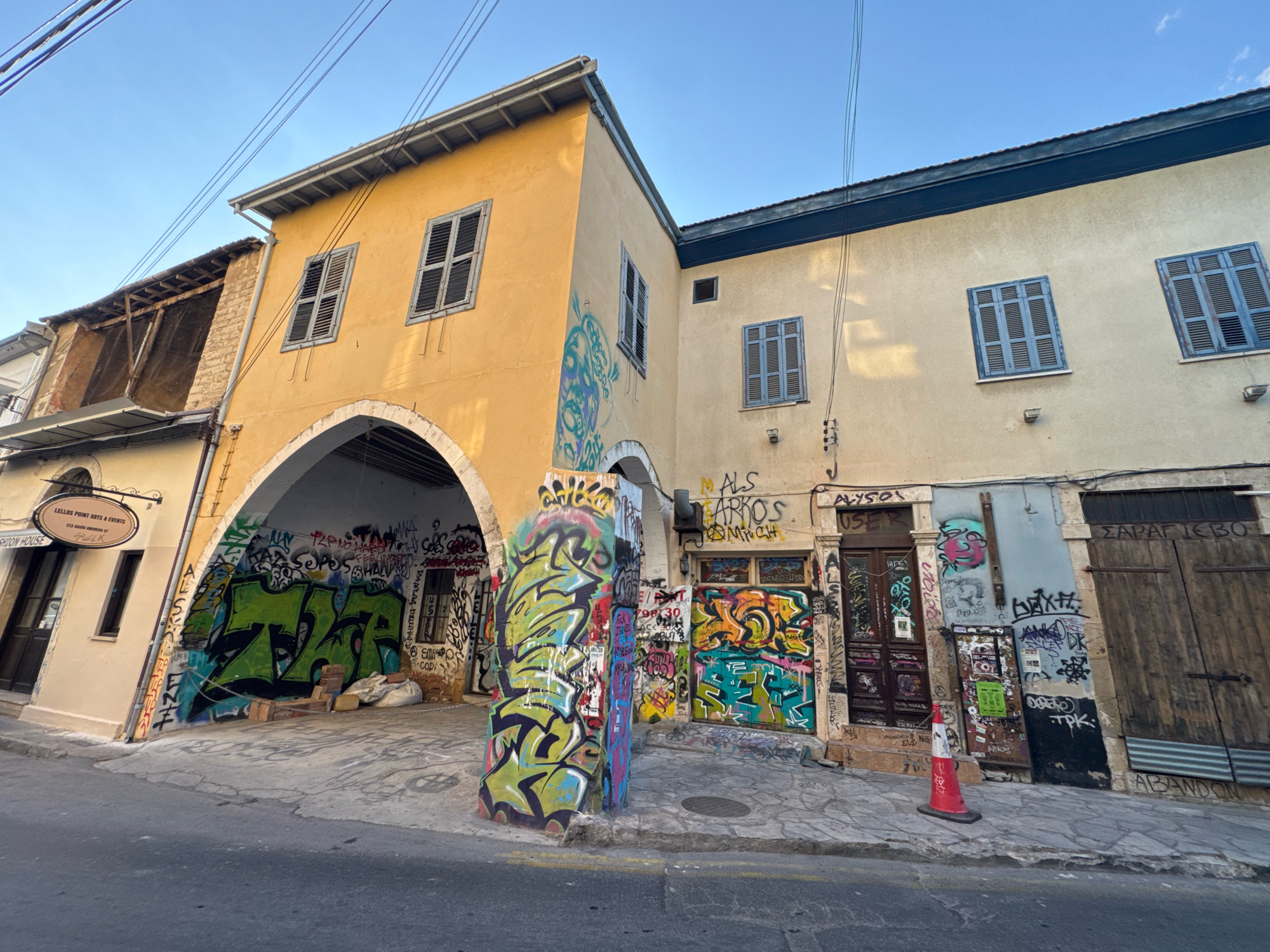 The image shows a street scene in Limassol, Cyprus, with a focus on two interconnected buildings covered in graffiti. The building on the left is a lighter yellow and has an arched entryway that is also heavily graffitied. Above the arch, there are two windows with dark shutters. To the left of this building, a narrower building is partially visible, with a sign that reads "LELLOS POINT ARTS & EVENTS" and "FASHION HOUSE". The building on the right is a pale yellow and has several windows with light blue shutters, as well as some smaller square windows. The street in front of the buildings is paved with stone and asphalt. A red and white traffic cone is standing on the sidewalk in front of one of the doors on the right. The overall impression is one of urban decay and artistic expression, with the vibrant graffiti contrasting with the weathered facades of the buildings.