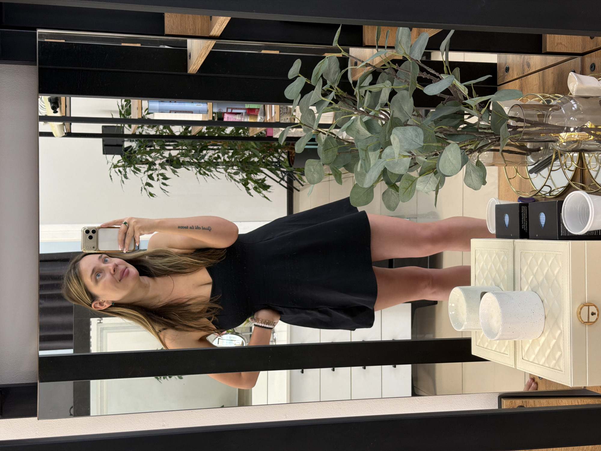 A woman is taking a mirror selfie in what appears to be a room with a large mirror. She is wearing a black dress and has long, blonde hair. Her left hand is on her hip, and she is holding a smartphone in her right hand. In the reflection, a plant is visible behind her, as well as shelves with various items. In the foreground of the mirror, there are two white candles on a decorative box. The background shows a white dresser with drawers. The scene is lit by indoor lighting. The location is not identifiable from the image itself.