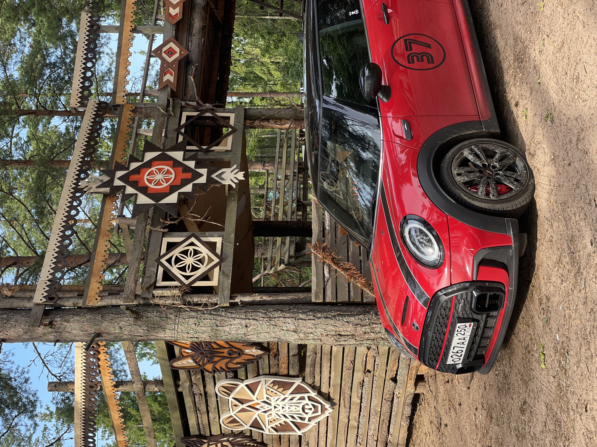 A red Mini Cooper is parked on a dirt road in front of a wooden structure decorated with geometric carvings and animal motifs. The Mini Cooper has black racing stripes on its hood and a black roof. A circular decal with the number "37" is visible on its side. The wooden structure appears to be a gate or entrance, with intricate carvings of animal faces and geometric patterns. The background consists of a forest with tall trees and blue sky. The license plate of the car reads "0267 AA 250" and indicates it is registered in Russia. The scene appears to be located in Ozerki (Krasnodolinskaya), Russia. There are no people visible in the image.
