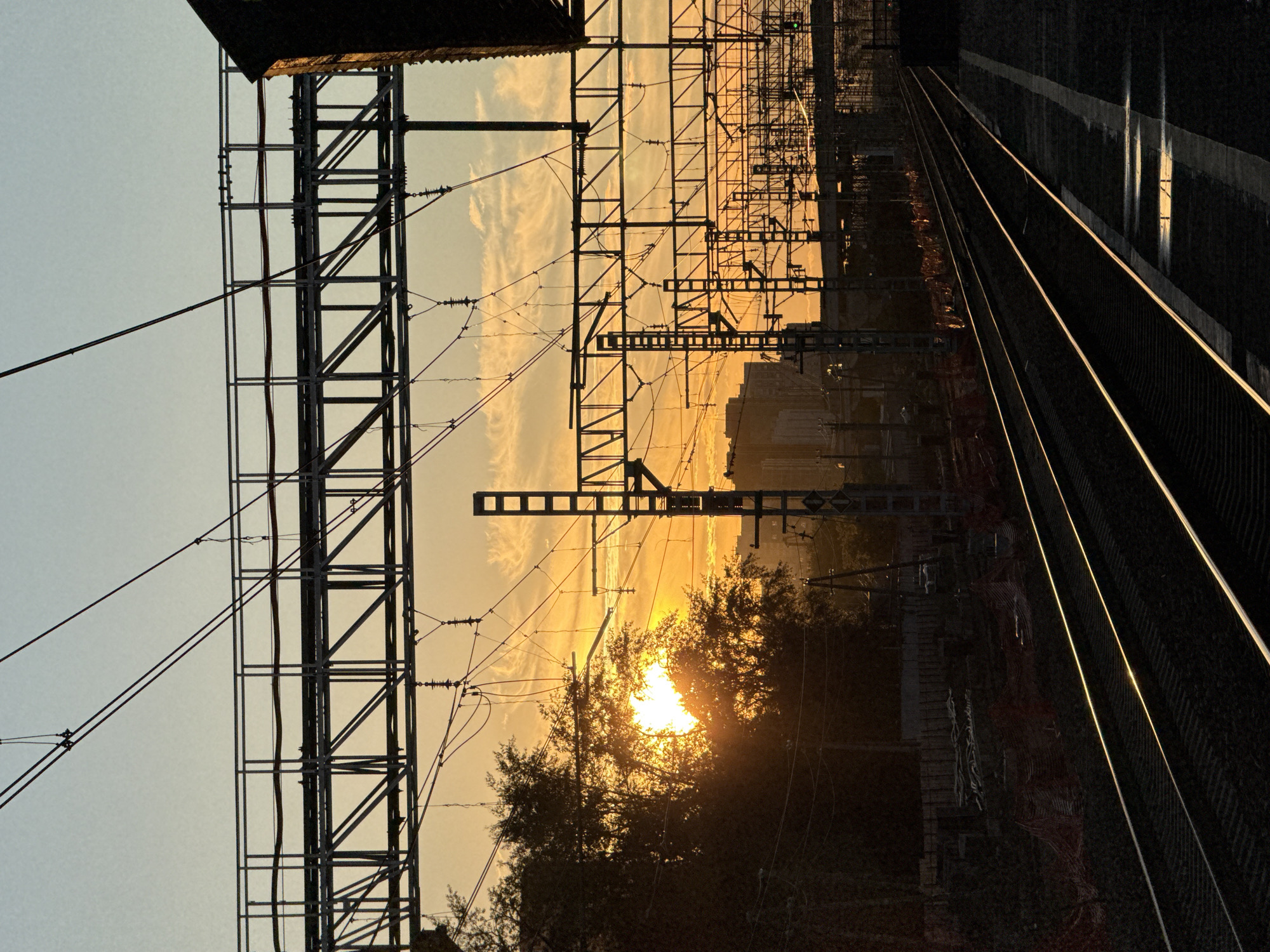 The image depicts a railway track stretching into the distance, bathed in the warm glow of a setting sun. Overhead, a complex network of power lines and support structures crisscrosses the sky, silhouetted against the orange and yellow hues of the sunset. Wispy clouds drift across the horizon, adding texture to the sky. In the foreground, trees with dark, dense foliage partially obscure the view of the sun. The railway tracks themselves are dark, with reflections of the sky visible on their surface. In the far distance, a city skyline with tall buildings can be seen. The overall mood is serene and atmospheric, with the sunset casting long shadows and creating a dramatic contrast between light and dark. The scene evokes a sense of a tranquil end to the day, with the industrial elements of the railway juxtaposed against the natural beauty of the sunset. The location is identified as Moscow, Russia. There is no visible text in the image.