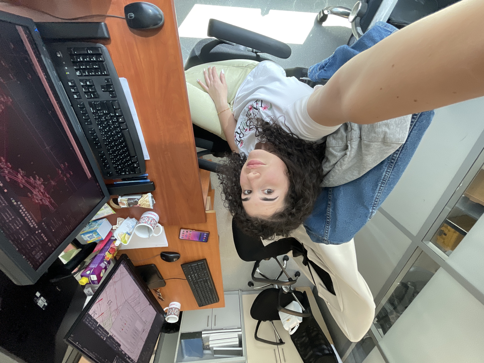 A selfie is taken from a high angle inside an office setting. A woman with curly dark hair is in the foreground, wearing a white t-shirt, a gray sweatshirt, and blue jeans. She is positioned in an office chair facing the camera. The office desk is on the left side of the frame. There are two computer monitors. One displays what appears to be technical diagrams and schematics, and another shows a map. There is a black keyboard and computer mouse on the desk. Miscellaneous objects are visible, including snacks, beverages, and cups. There is a mobile phone on the desk. In the background, there is another office chair and a window with a view of an office building. The location is plausibly Sofia, Bulgaria based on the Eastern European appearance of the woman and the general office aesthetic.