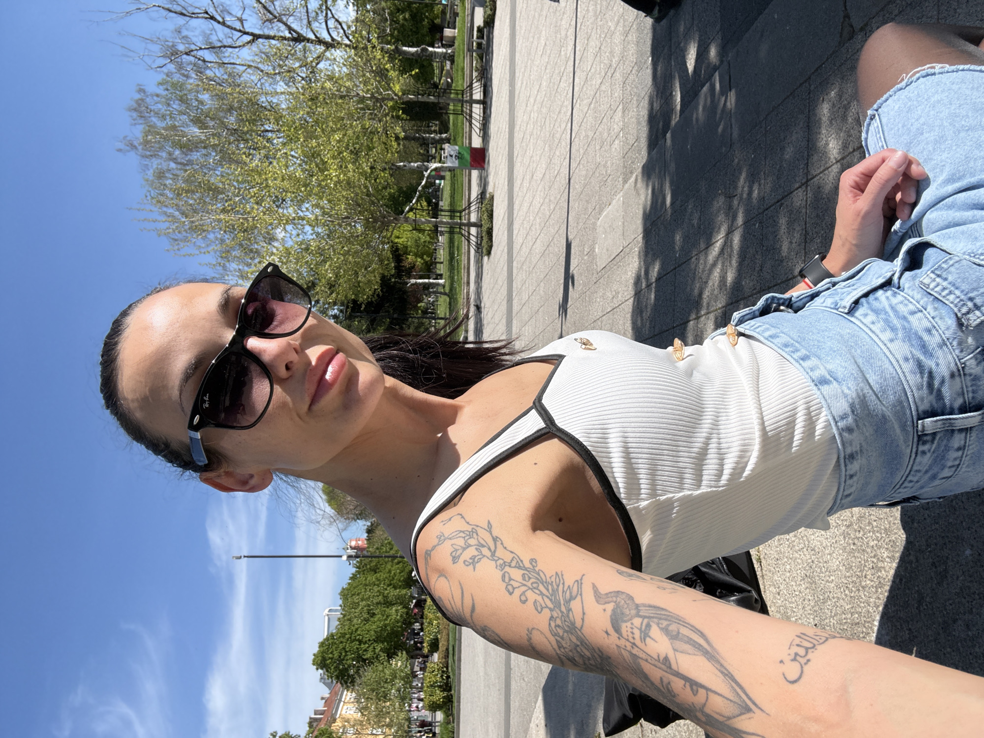 A young adult female is featured in a vertical outdoor selfie, taken on a bright, sunny day in what appears to be a park or public square in Pleven, Bulgaria. Strong shadows suggest it is midday or early afternoon. The woman is in the foreground, facing the camera with a neutral to slightly smiling expression, her lips slightly parted. She wears dark sunglasses with "Ray-Ban" visible on the side. Her dark hair is pulled back, and she is dressed in a white ribbed tank top with black trim and light-wash denim shorts. A black smartwatch is visible on her left wrist.
Her arms are adorned with tattoos; intricate floral and bird designs cover parts of her arms, and on her right forearm, Arabic script is clearly visible, which spells "فلسطين" (Palestine).
The background features a clear blue sky with minimal clouds. Beneath the sky, numerous green trees with lush foliage indicate spring or early summer. Paved pathways and grassy areas characterize the park setting. In the mid-ground, among the trees, a vertical banner or flag with green, white, and red stripes is partially visible. Buildings can be seen in the far distance, providing an urban backdrop to the serene park environment. The overall scene captures a relaxed moment outdoors.