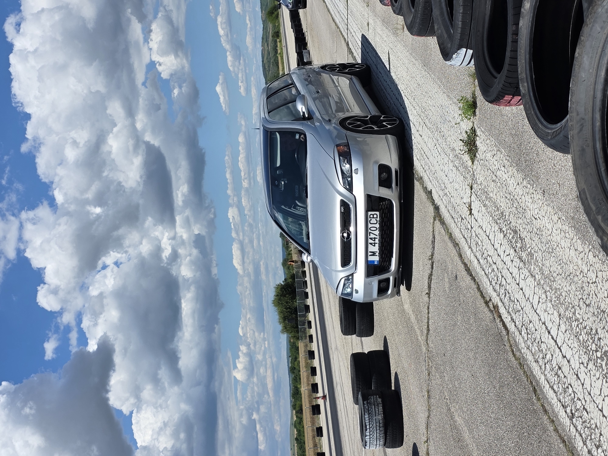 A silver station wagon is parked on an asphalt surface. The car has a Bulgarian license plate that reads "M 4470 CB". Several stacks of tires are visible on the left and right sides of the image. The background shows a flat, rural landscape with some trees and hills under a partly cloudy sky. This scene is located on a racetrack in Akatsievo, Bulgaria.