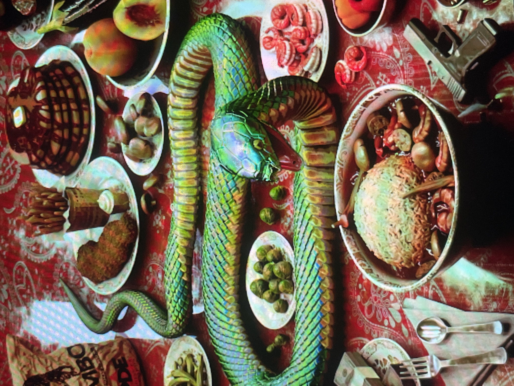 The image presents an elaborate, overhead view of a surreal table setting, dominated by a large, coiled snake with shimmering green and blue scales. The snake's head, featuring an open mouth and visible fangs, is positioned centrally amidst a variety of dishes. The table is covered with a red tablecloth adorned with a white paisley pattern.
Surrounding the snake are numerous plates and bowls filled with food. These include a dark, fluted dish, peaches (some halved), small light-colored items, shrimp and possibly sausages on a white plate, small green vegetables resembling Brussels sprouts, and green beans. A large bowl contains rice mixed with other ingredients and a sauce. Another dish holds brown, patty-like food, accompanied by fries or crackers and a dip.
Contrasting sharply with the culinary display, a silver/grey handgun is placed on the tablecloth in the upper right, alongside what appears to be a bullet. In the lower right, stacks of US dollar bills are arranged on a small plate next to cutlery, including forks and spoons. A piece of cloth or bag in the bottom left corner partially displays the text "MBO DE".
The scene is lit dramatically, creating strong highlights on the metallic-looking snake and pronounced shadows, enhancing the textures of the food and tablecloth. No people are visible. The overall impression is that of a symbolic or artistic arrangement rather than a literal meal. This media file originates from Sofia, Bulgaria.