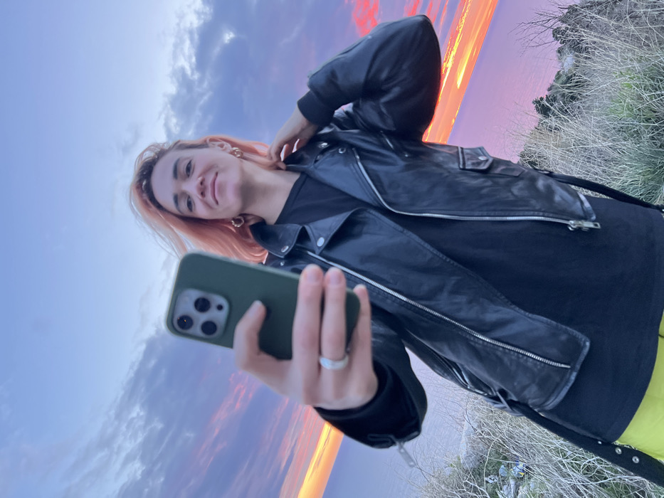 A young woman, angled towards the camera, holds a green-cased smartphone to take a selfie. She's wearing a black t-shirt and a leather jacket, and her hair is dyed a light pinkish hue. Behind her, the sky is a vibrant mix of colors from a sunset, transitioning from blue at the bottom to orange and pink higher up. The scene has a slight tilt, and some grassy foliage is visible in the lower right corner.