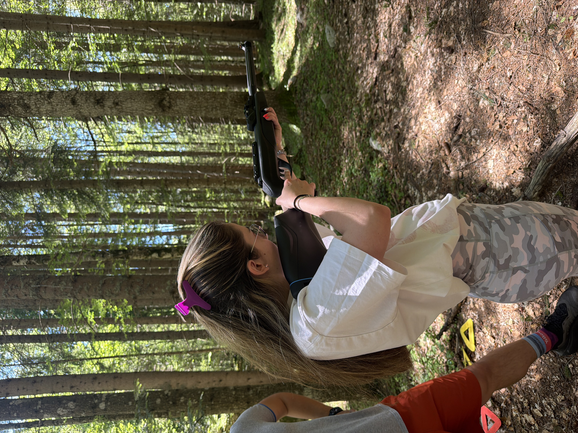 The image depicts a young woman holding a rifle in a forest environment. She is wearing glasses, a white t-shirt, and camouflage leggings, with a pink hair clip in her long brown hair. Her right hand is on the trigger of the rifle, and her left hand is supporting the barrel. Another person, partially visible on the left, is wearing red shorts and a grey t-shirt. The background consists of numerous tall trees, characteristic of a forest. The ground is covered with pine needles and fallen leaves. Several colorful triangular targets are visible on the forest floor. The lighting suggests it is daytime with sunlight filtering through the trees. The scene appears to be an outdoor recreational activity, possibly target practice, taking place in a wooded area.