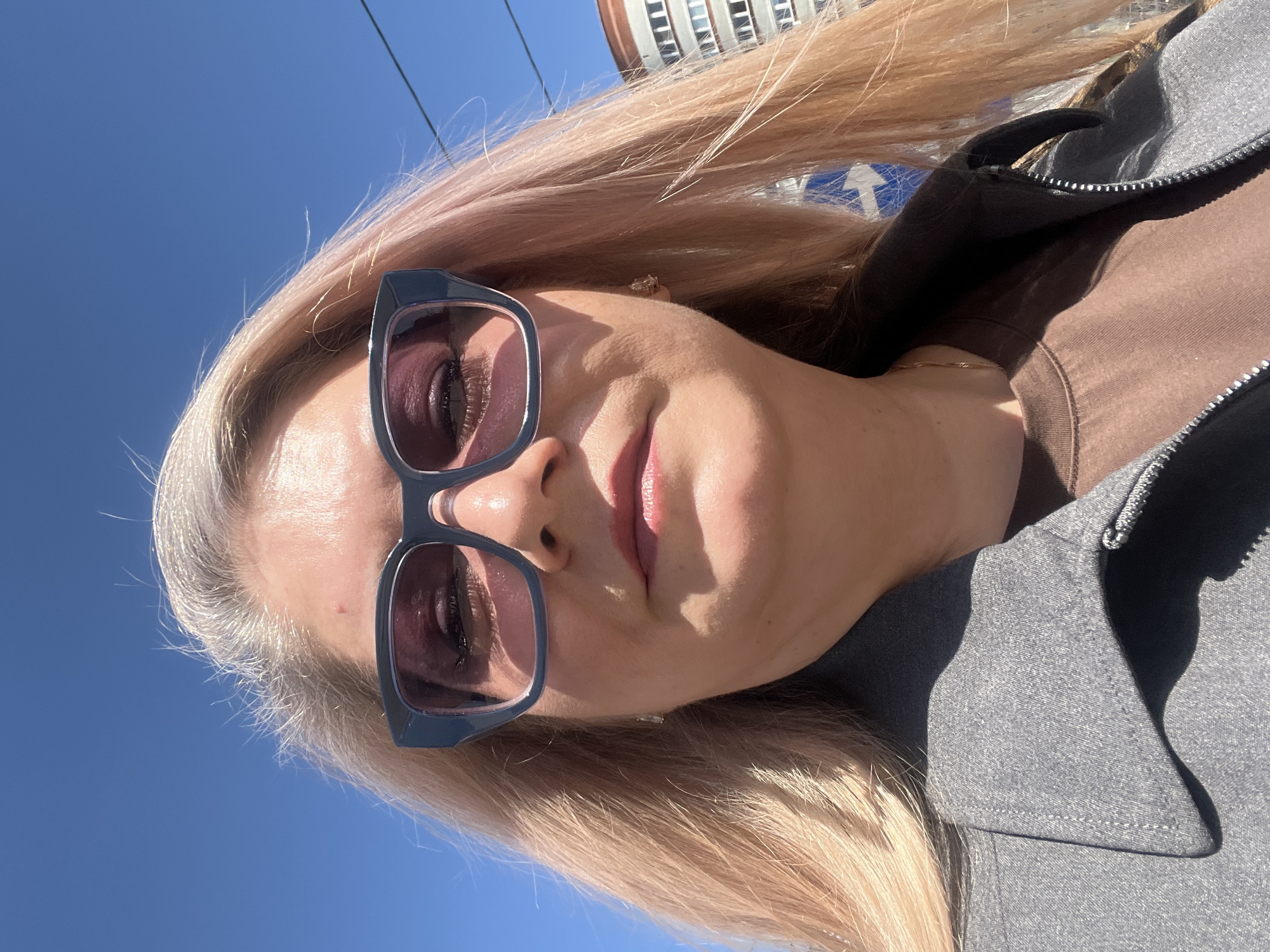 A selfie of a person with pinkish-blonde hair and sunglasses is tilted upward towards a blue sky. A building is partially visible in the upper left corner. The person is wearing a gray and brown jacket. This image may have been taken in Izhevsk, Russia.