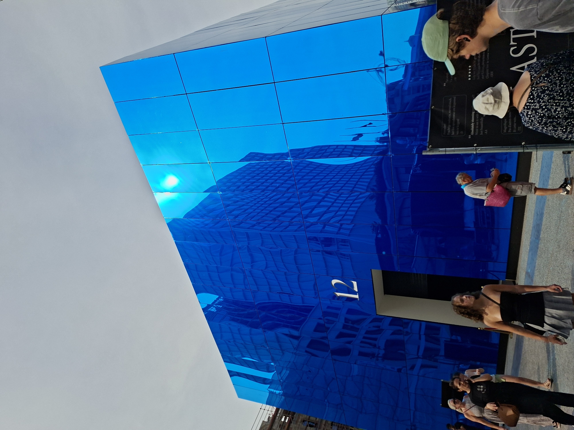 The image depicts the exterior of a modern building with a striking blue, reflective facade. The building, identified by the prominent number "12" above an entrance, is situated in a public space with several people in the foreground. The blue facade reflects distorted images of surrounding buildings and the sky, with the sun clearly visible as a bright spot on the upper right portion of the structure.
Several individuals are present: a woman in a black strapless top and grey skirt is walking to the right; two women and another person are walking towards the left; a man in a light-colored shirt and shorts is standing and looking at his phone, carrying a pink bag; and two young men, one wearing a green baseball cap, are looking at a black sign to the right. The sign appears to be an information board or exhibit with text and possibly ratings.
The environment suggests an urban setting, likely a street or plaza, given the presence of pedestrians and the architectural style of the building and reflected structures. Construction scaffolding is visible in the background on the left, indicating ongoing development or renovation in the area. The overall impression is one of contemporary architecture and public engagement with art or a notable building. The location is identified as Brussels, Belgium.