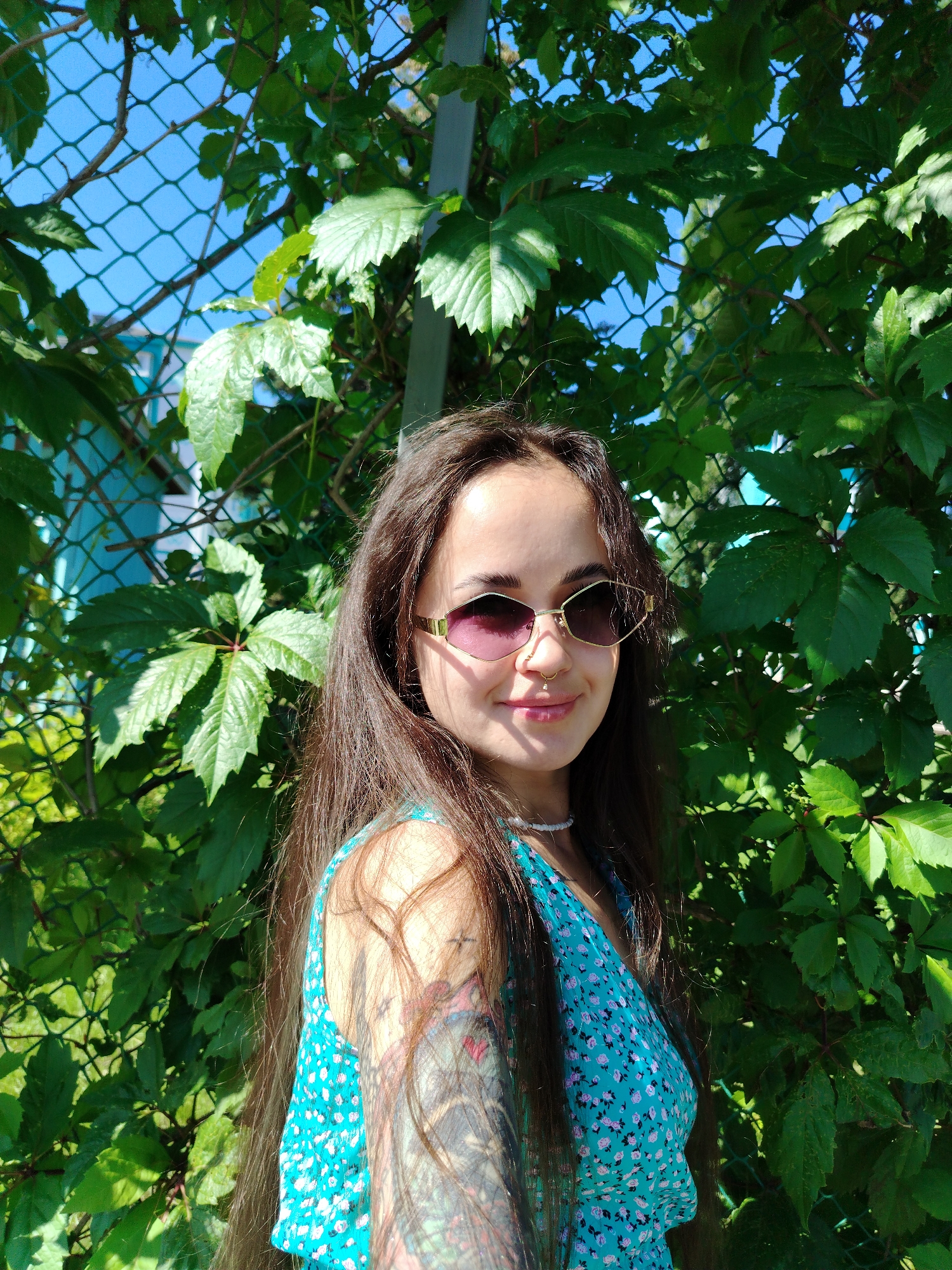 A young woman with long brown hair is standing outdoors in front of a green leafy vine and a chain-link fence. She is wearing sunglasses with gold frames and a light blue dress with a floral pattern. She has a nose ring and a necklace. Tattoos are visible on her arm. The background shows a bright blue sky and more greenery. The location is Yanaul, Russia.