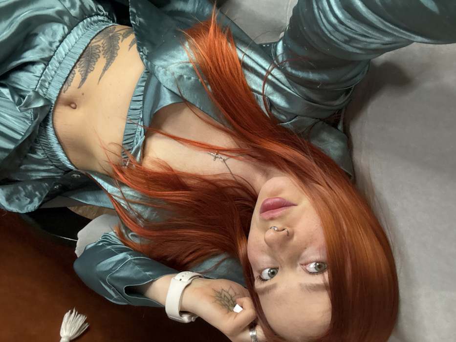 The image portrays a young adult female subject in an indoor setting in Rostov-on-Don, Russia. The subject has long, bright orange-red hair and green eyes, and is lying in a reclined position, looking directly at the camera. She is wearing a two-piece outfit of teal, satin-like material, consisting of a cropped long-sleeved top and matching bottoms, exposing her midriff. A silver-colored cross pendant hangs from a chain around her neck. Her left hand displays a small, circular tattoo resembling a lotus flower on the back, below a white smartwatch on her wrist and a silver ring on her index finger. Her fingernails are painted white. A large tattoo featuring multiple dark-inked fern or feather-like leaves is visible on her left rib cage and abdominal area. She has a septum piercing in her nose and a circular barbell piercing in her navel. The background is blurred, showing a dark reddish-brown object and a white tassel-like textile item on the upper left, with the subject resting on a light gray fabric surface.