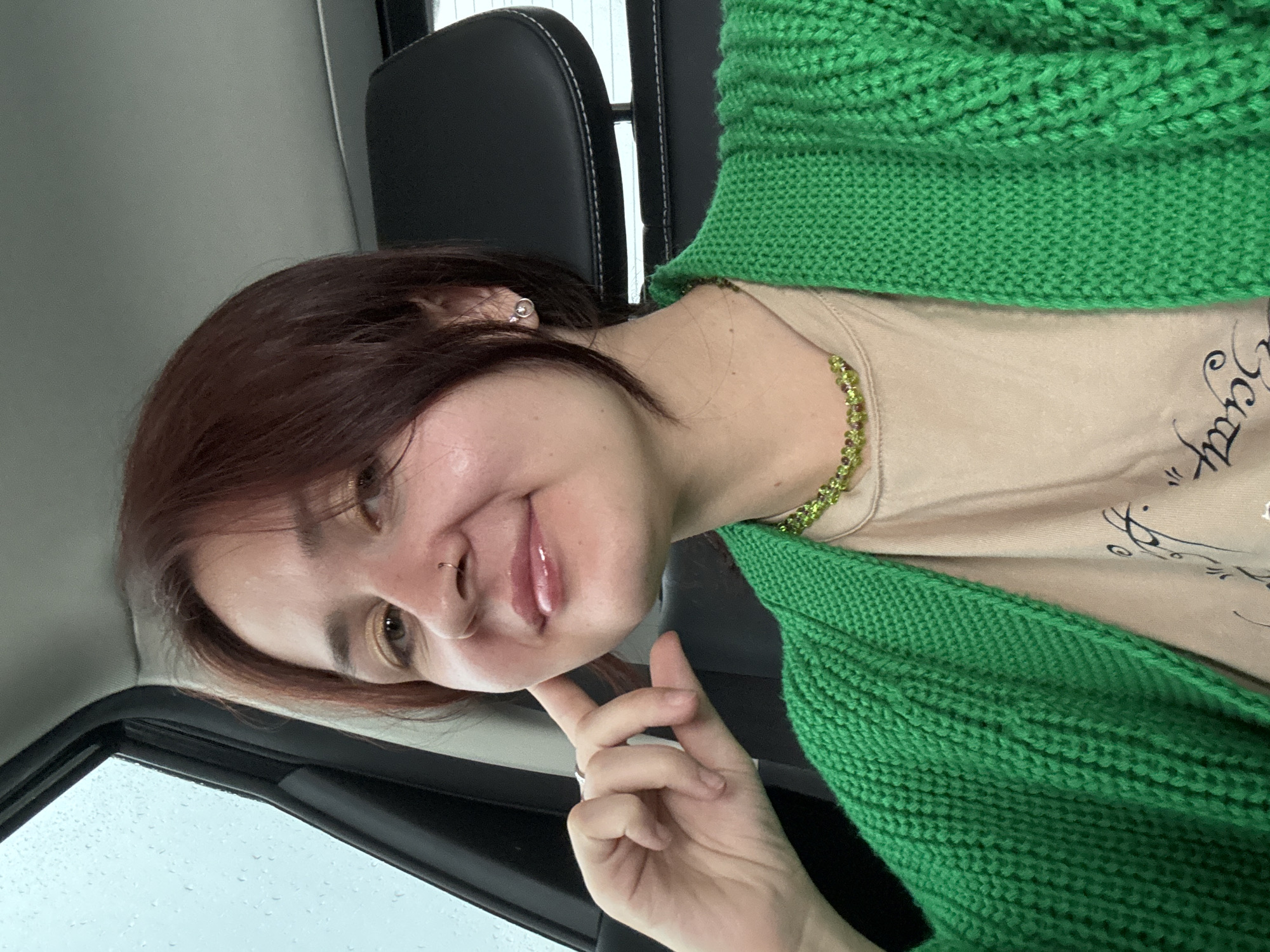 A young woman with auburn hair is sitting in the passenger seat of a car. She is wearing a beige t-shirt with a black graphic print and a bright green chunky knit cardigan. She has a nose ring and a silver earring in her left ear. A green beaded necklace is visible around her neck. Her left hand is resting on her chin, and she is looking towards the camera with a slight smile. The window to her left shows a blurry, overcast sky with raindrops. The interior of the car, including the headrest, is visible in the background. This image was taken in Naberezhnye Chelny, Russia.