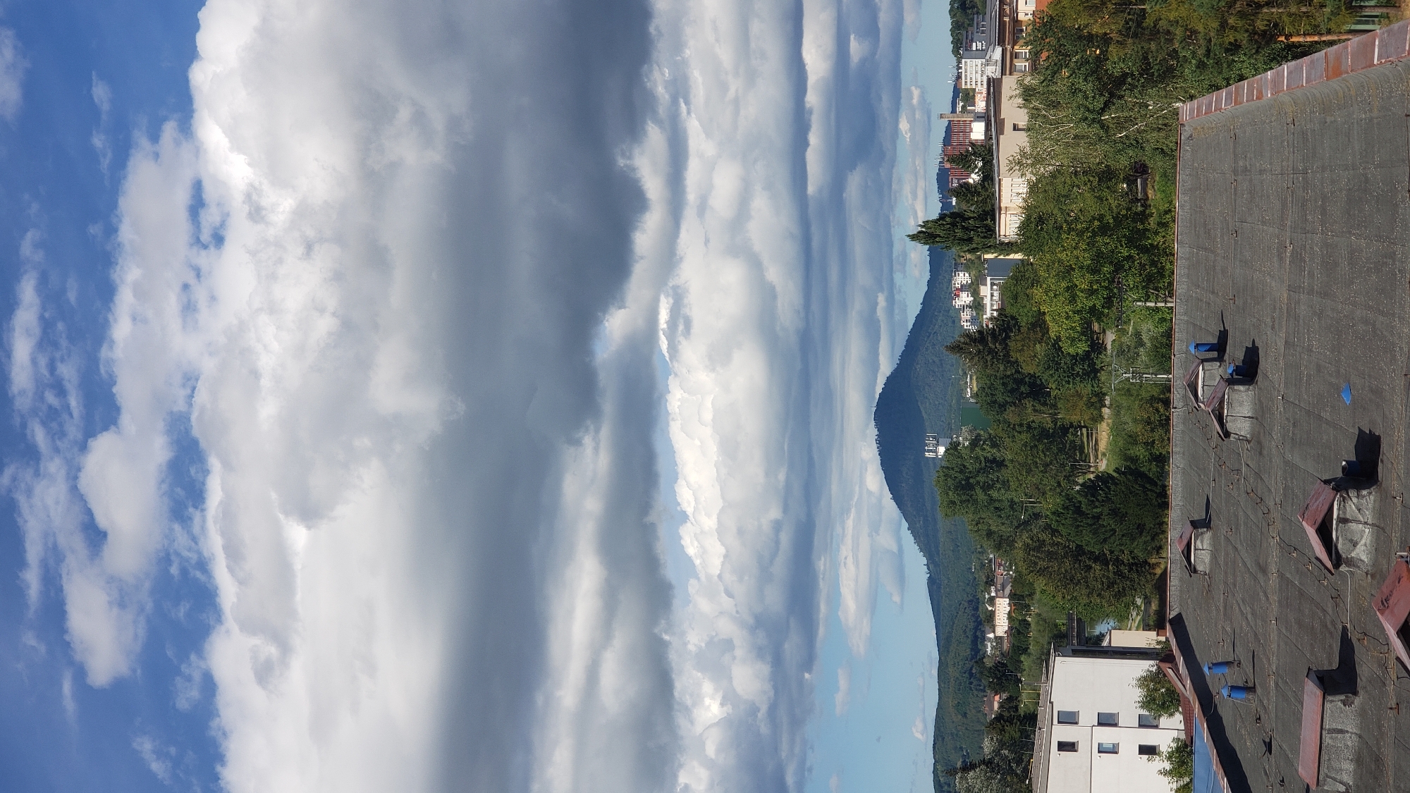 The image shows a panoramic view of a cityscape with a prominent mountain in the background. The foreground is dominated by the flat, dark, and weathered roof of a building, equipped with several skylights and ventilation pipes. Beyond the roof, a residential building with white and blue elements is visible. Further into the scene, lush green trees form a dense line, behind which a body of water can be glimpsed. In the middle ground, the city unfolds with buildings of various architectural styles and heights, including what appears to be a sports field or park. The backdrop is defined by a large, verdant mountain, on which a telecommunications tower is situated. The sky is a dynamic mix of bright blue patches and heavy, grey-to-white clouds, suggesting a changeable weather pattern. This scene is likely from Prešov, Slovakia, given the distinctive mountain shape and urban layout.