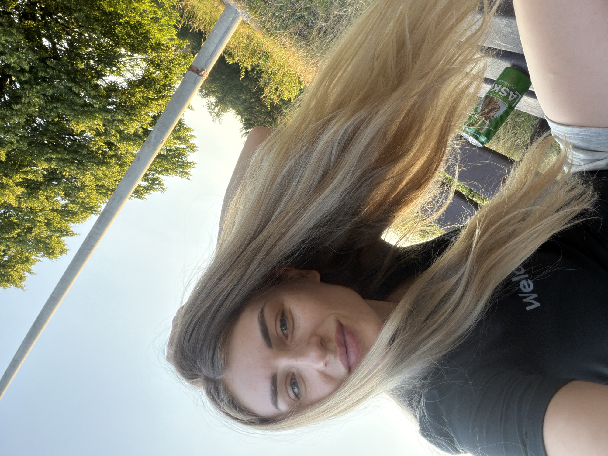 A woman with long blonde hair is pictured in a selfie taken outdoors. She is wearing a dark t-shirt and grey shorts. Her left arm is resting on her head, and she is smiling at the camera. Next to her, on a wooden bench, is a green can of Lasko Zlatnog beer. The background shows green trees and a clear blue sky, suggesting it is daytime and possibly a park or a natural area. A grey pole is also visible in the right side of the image. The lighting indicates the sun is low, possibly late afternoon or early morning.