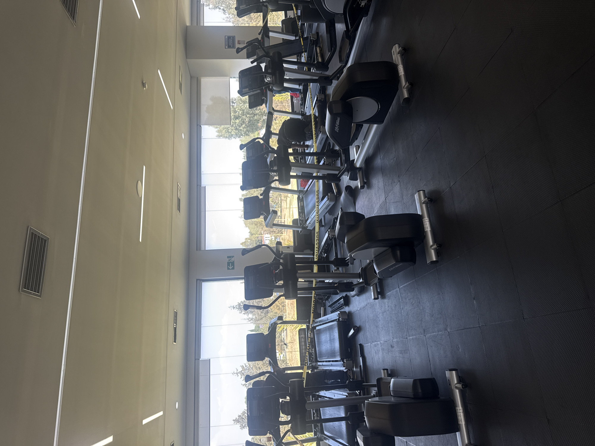 The image depicts a fitness center or gym in San Andrés Cholula, Mexico. The gym features multiple treadmills and stationary bikes lined up in rows. Yellow caution tape is wrapped around some of the equipment, indicating they are out of service. Large windows line one side of the gym, offering a view of trees and foliage outside. The floor is covered with black rubber mats. The walls are light-colored and have built-in linear light fixtures.