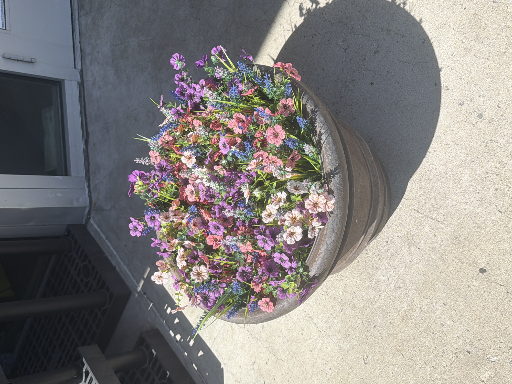 A flower-filled planter sits on a concrete surface outside a building. The planter is round and made of a dark, textured material, possibly stone or concrete. It is overflowing with artificial flowers in various shades of pink, purple, and blue, mixed with greenery. In the background, a metal grate is visible, along with dark vertical posts, suggesting an entrance or a covered area. The building facade is light-colored, with a glass window and a door. Sunlight casts a shadow of the planter onto the concrete.