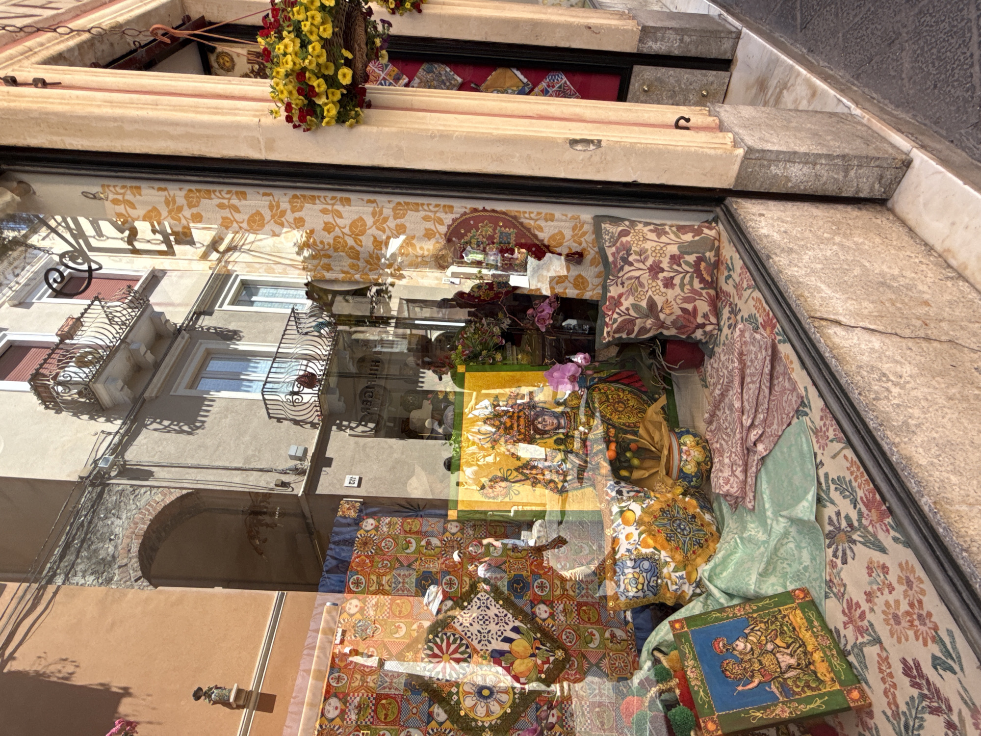The image shows the reflection of a building with balconies in the window of a store. Above the window, there's a flower basket hanging. Inside the store, there are patterned textiles and decorative objects on display. The reflection suggests the store is located in a city with old architecture.