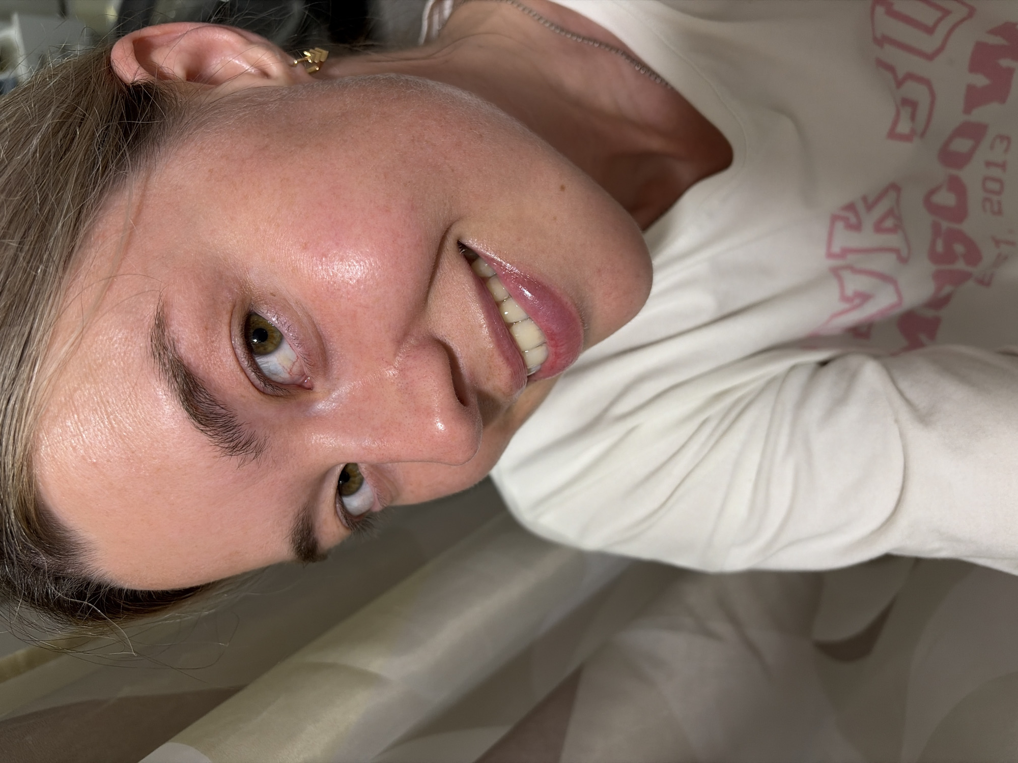 A close-up portrait of a young woman with blonde hair and green eyes, taken from a slightly upward angle. She is wearing a white long-sleeved t-shirt with pink lettering that reads "PINK SOLO MOSCOW EST. 2013". The woman has clear, glowing skin and is smiling, revealing her teeth. She is wearing a small, gold heart-shaped earring on her right ear and a silver chain necklace. The background consists of a beige and white striped shower curtain. The image was taken in Chaikovsky, Russia.