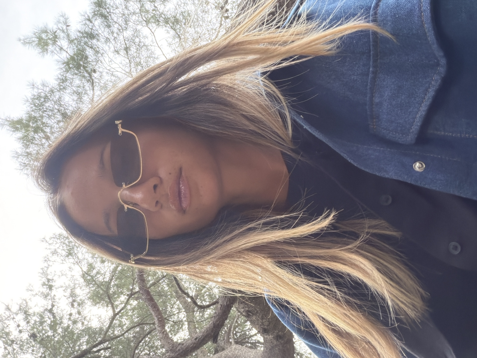 A woman wearing sunglasses and a dark blue denim jacket is pictured from a low angle, with trees and a bright sky visible in the background. The location is Unknown, Unknown.